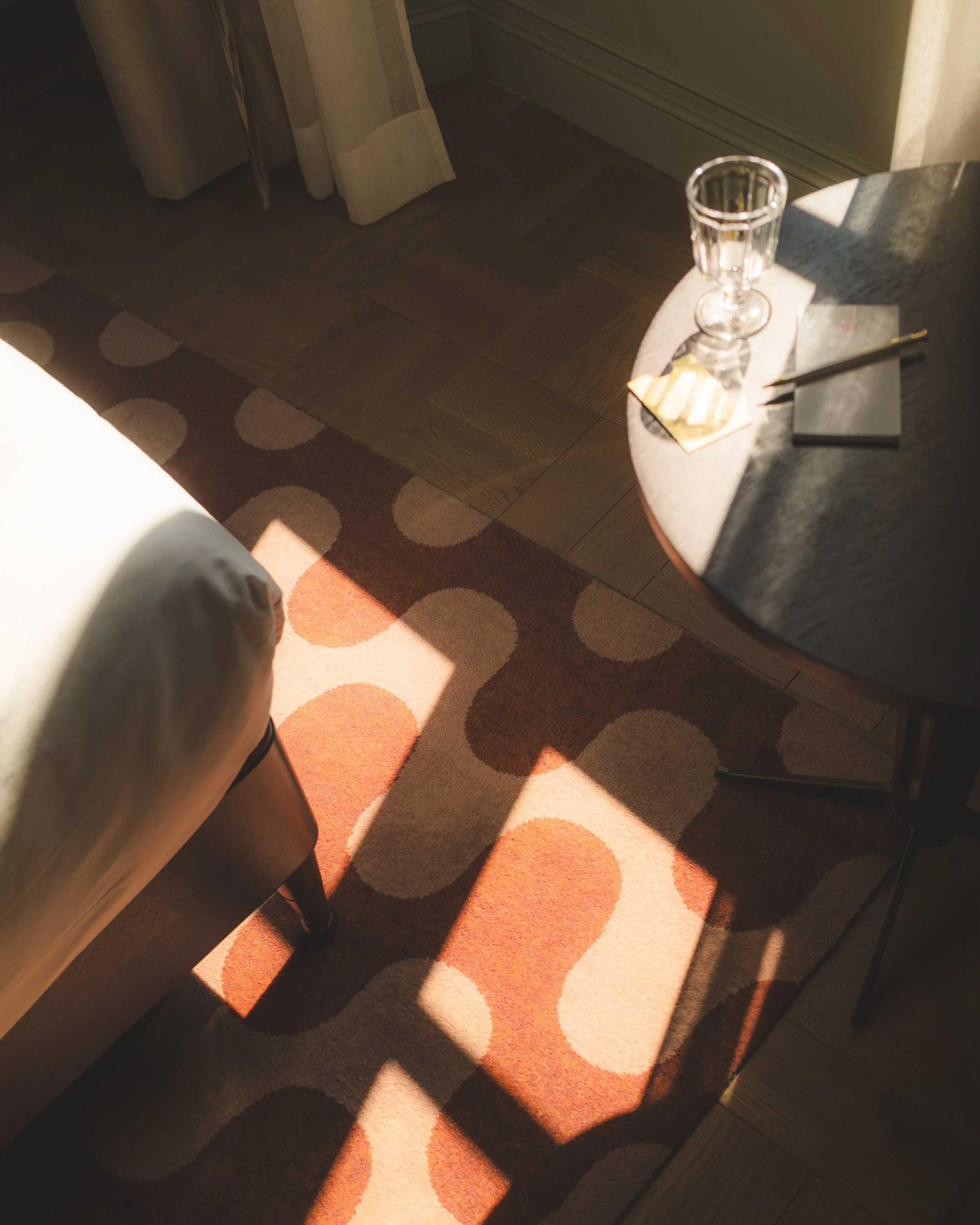 A corner of a room with wooden flooring, a patterned rug, a small round table with books, a glass, and a pen, partially illuminated by sunlight.