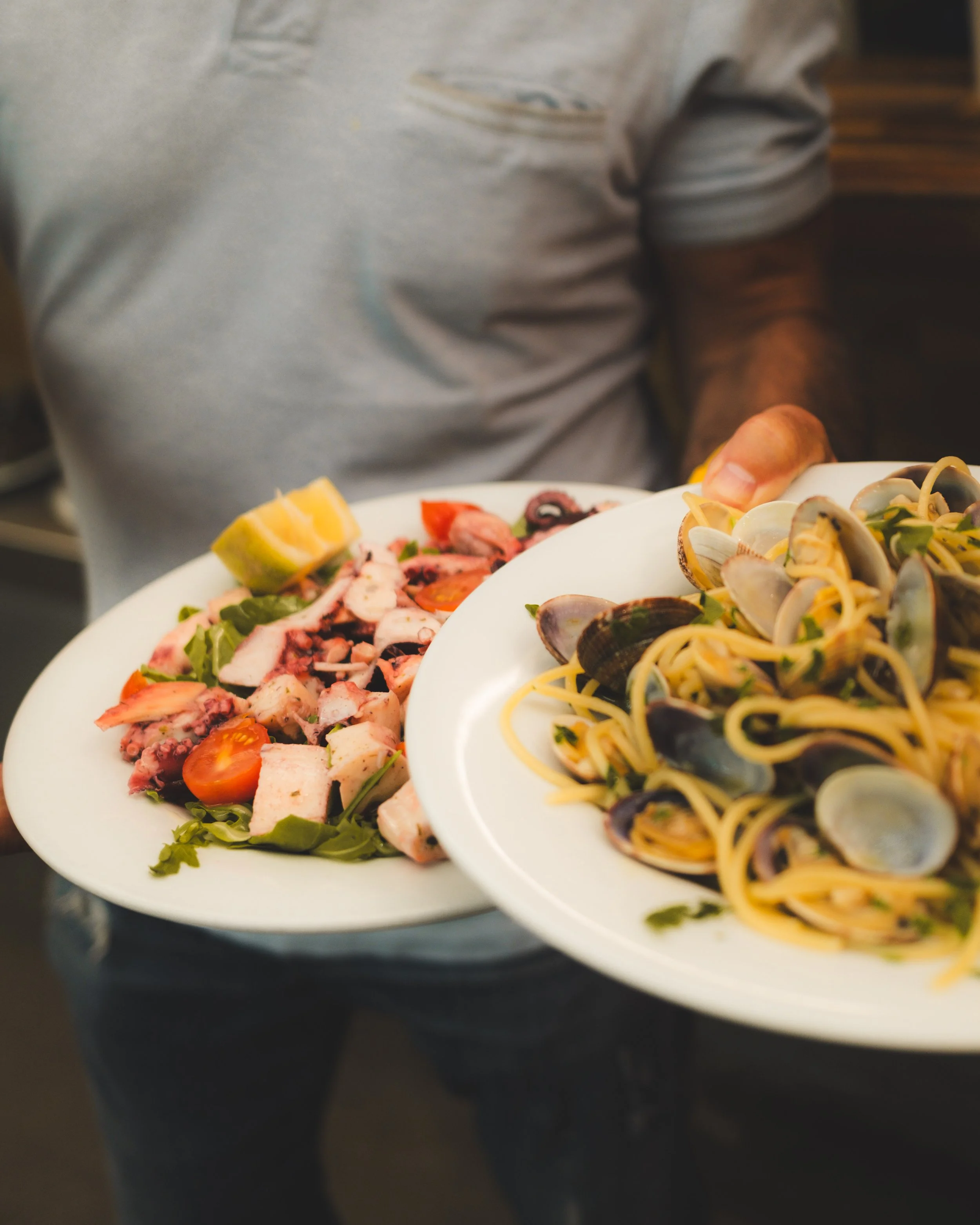 Person holding two plates of Italian food, one with seafood pasta and the other with a salad with tomatoes and grilled octopus. Lionel Chu - Hotel & Travel Photographer