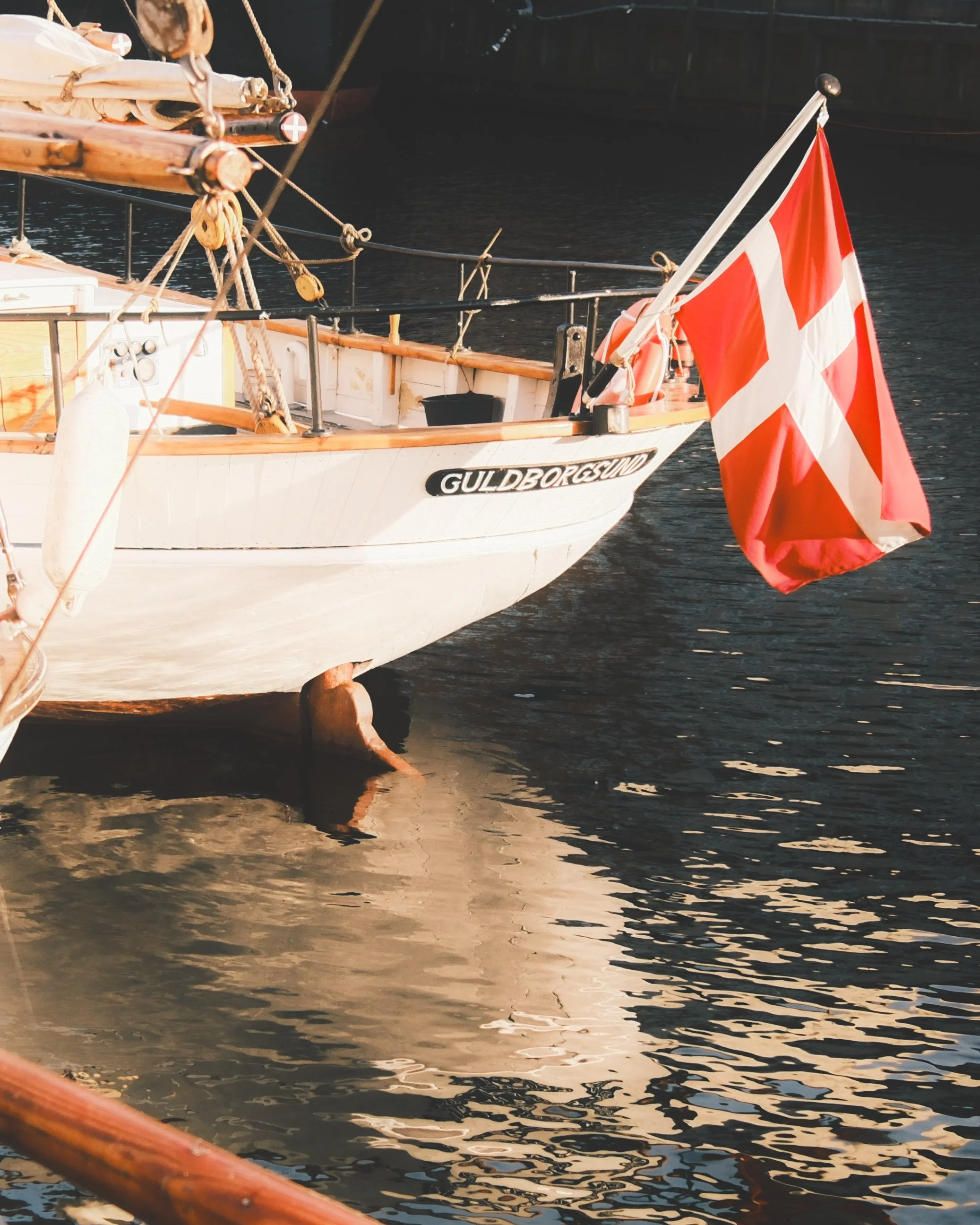 Close-up of a boat with a Danish flag, moored in the water, with ropes and wooden details visible.