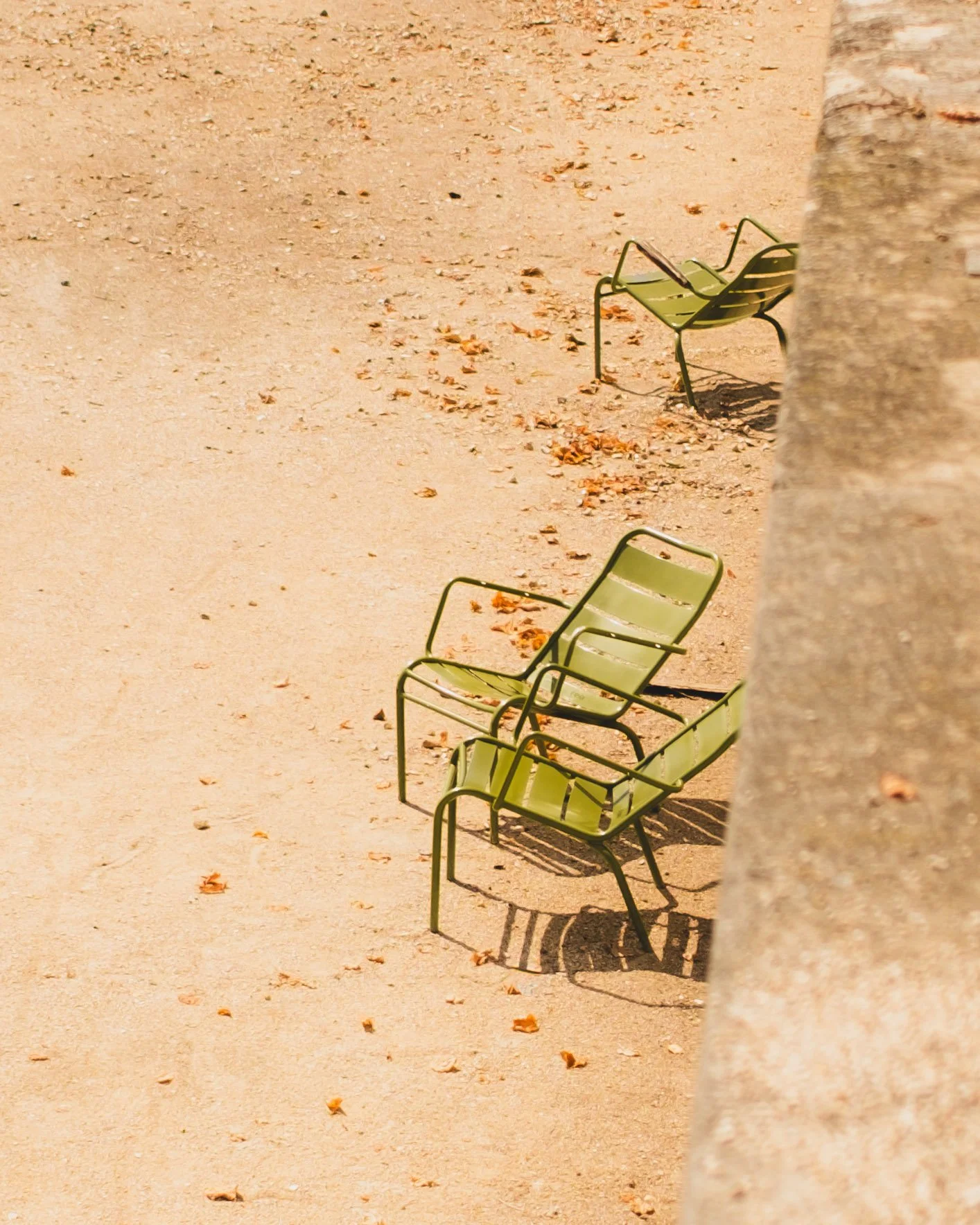 Three empty green chairs on brown sandy ground with long shadows and a blurred stone foreground. Jardin des Tuileries, Paris. Lionel Chu - Hotel & Travel Photographer.