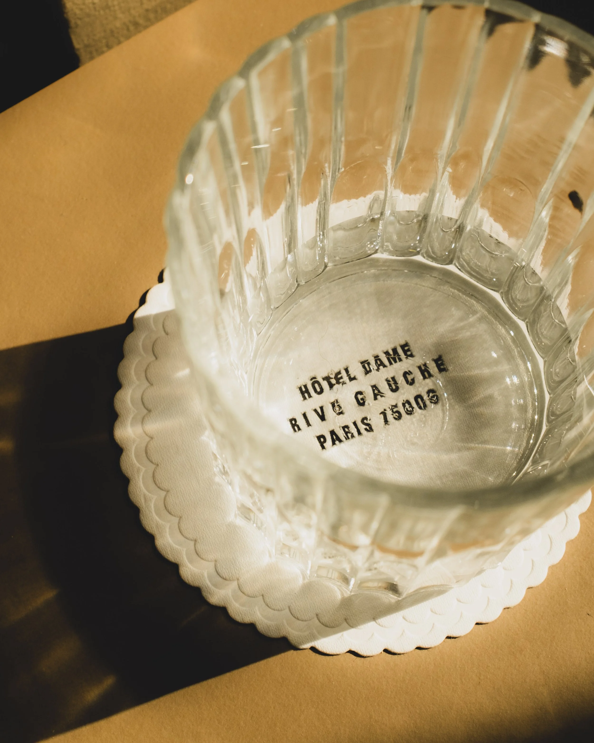 Clear glass cup with engraved text on the bottom and a scalloped edge, placed on a white paper doily on a wooden surface.