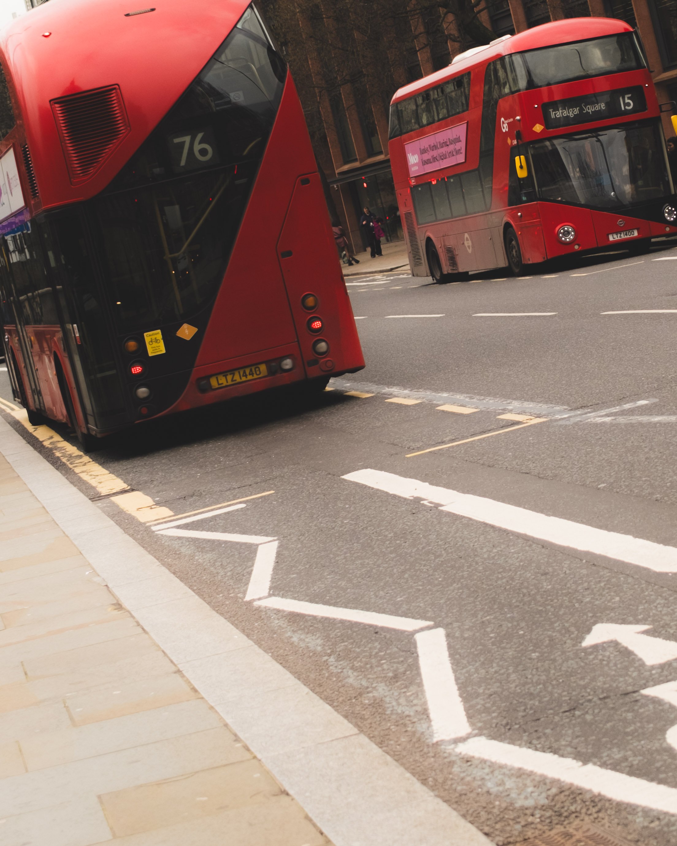 Two red double-decker buses on a city street, one with route 76 and the other with route 15, near a sidewalk with a few pedestrians.