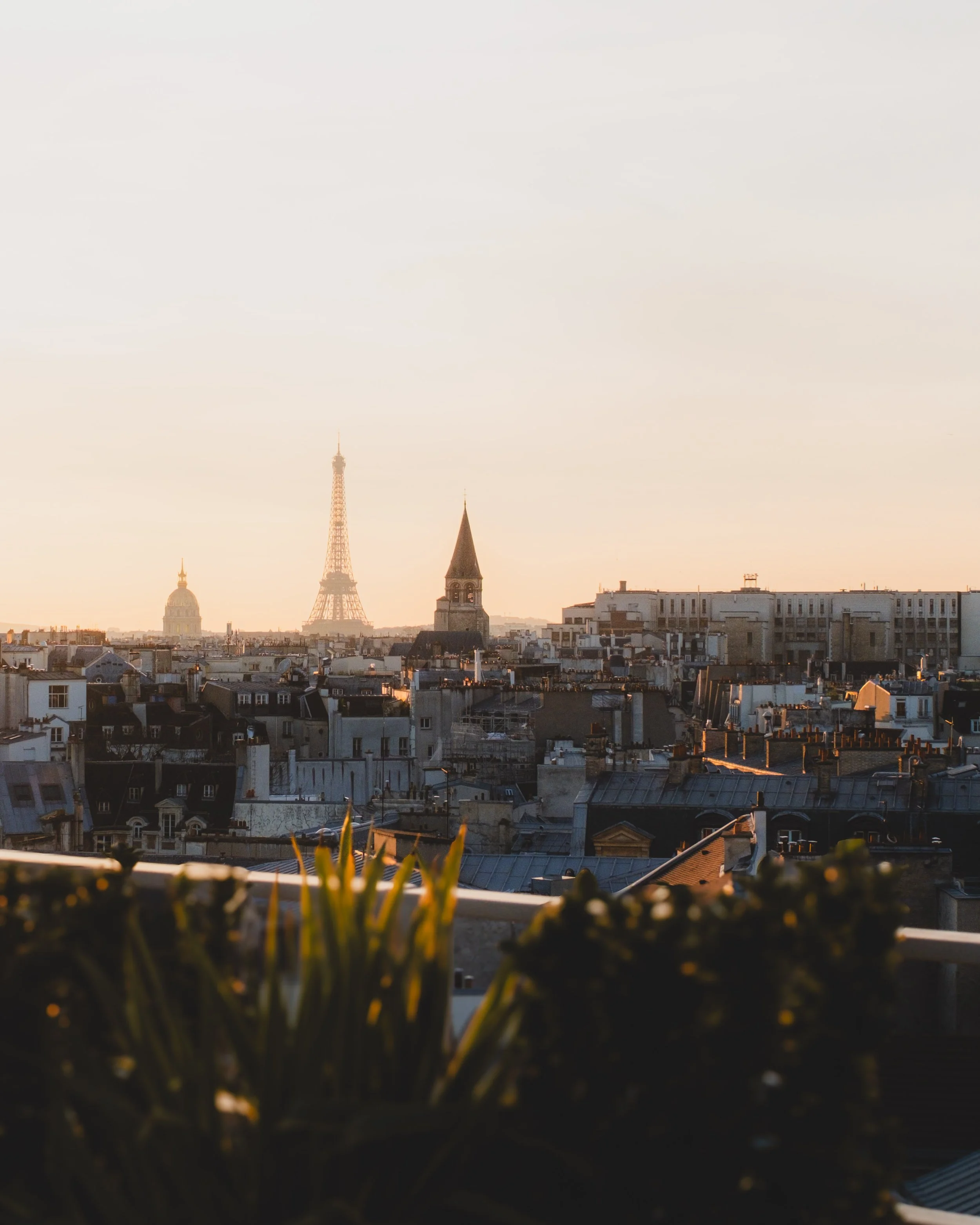 Paris cityscape at sunset with the Eiffel Tower, a church with a steeple, and rooftops visible in foreground.