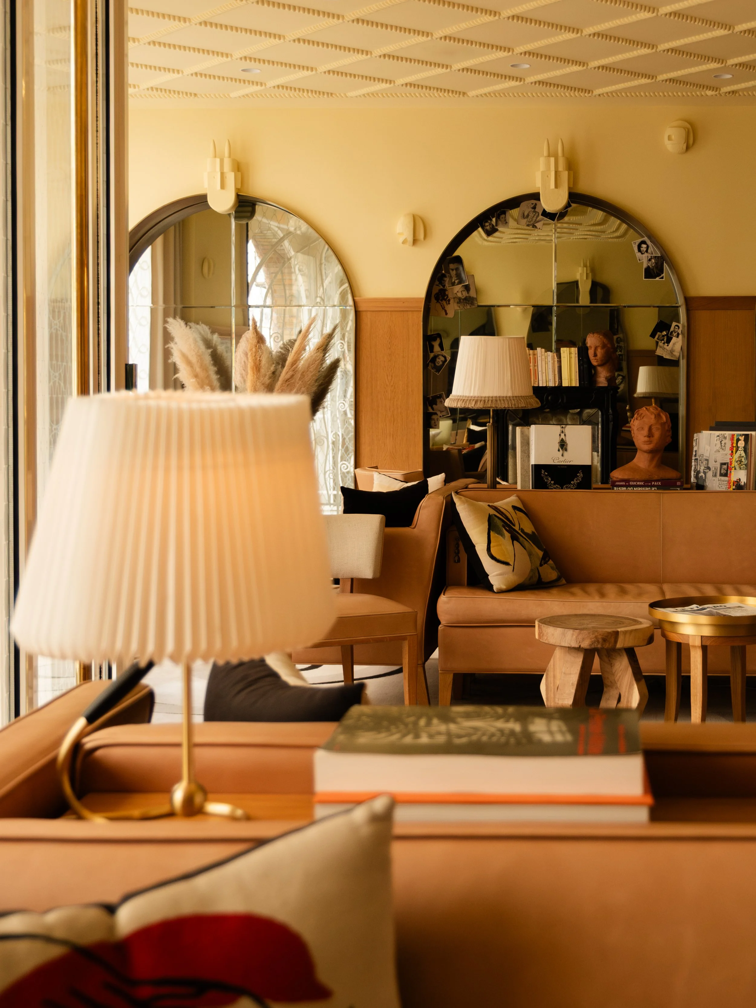 A cozy interior hotel lobby with beige sofas, wooden tables, a lamp, and decorative pillows. A mirror on the wall reflects books and sculptures, with natural light coming through a window. Lionel Chu - Hotel & Travel Photography