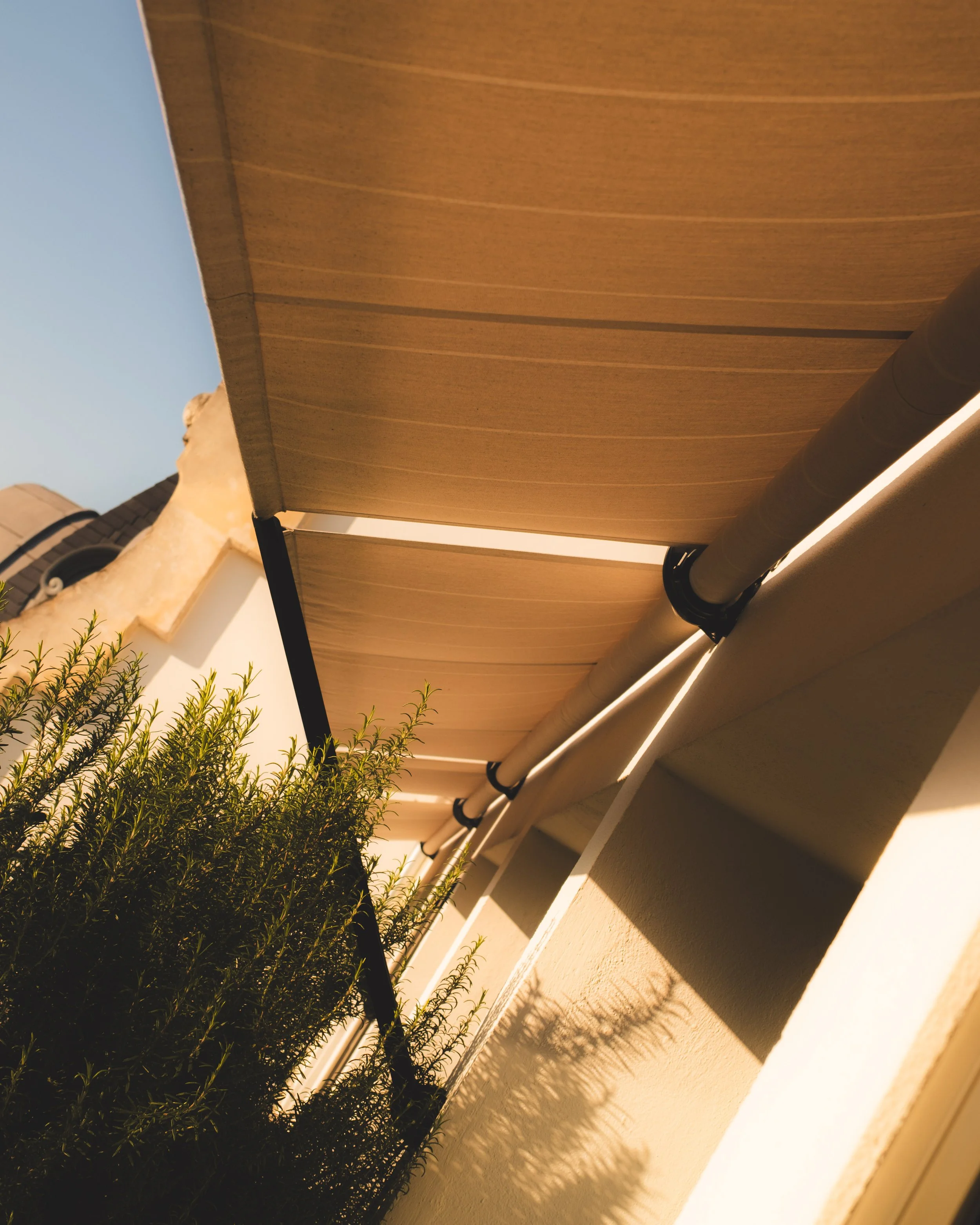 A view of a house exterior showing a beige wall with a shadow of a plant, a roof with grey tiles, a white soffit, and a brown gutter pipe, taken during sunlight with clear sky.