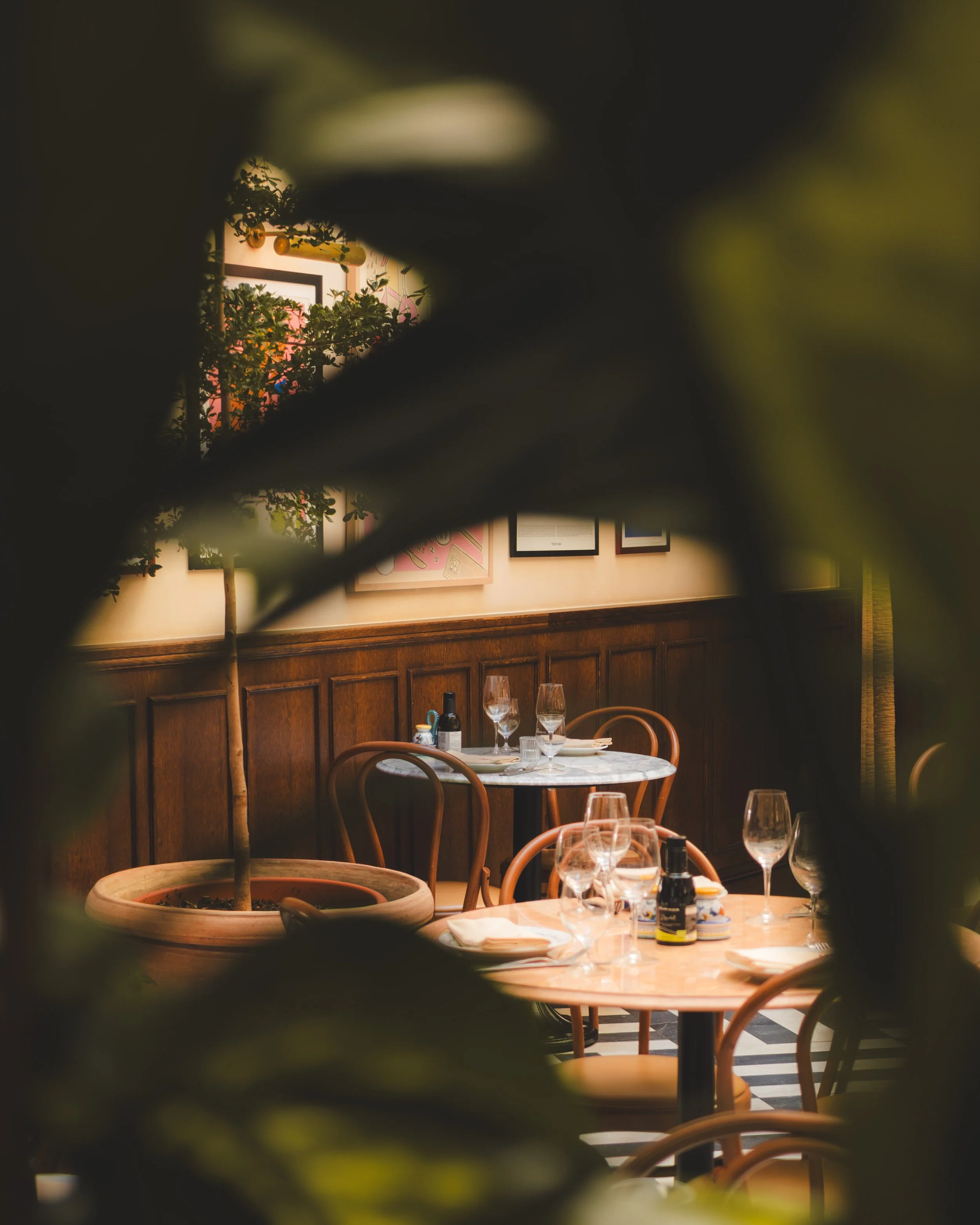 A view of a restaurant interior through a small gap in the foreground, showing tables set with wine glasses, bottles, and tableware, with framed artwork on the wall.