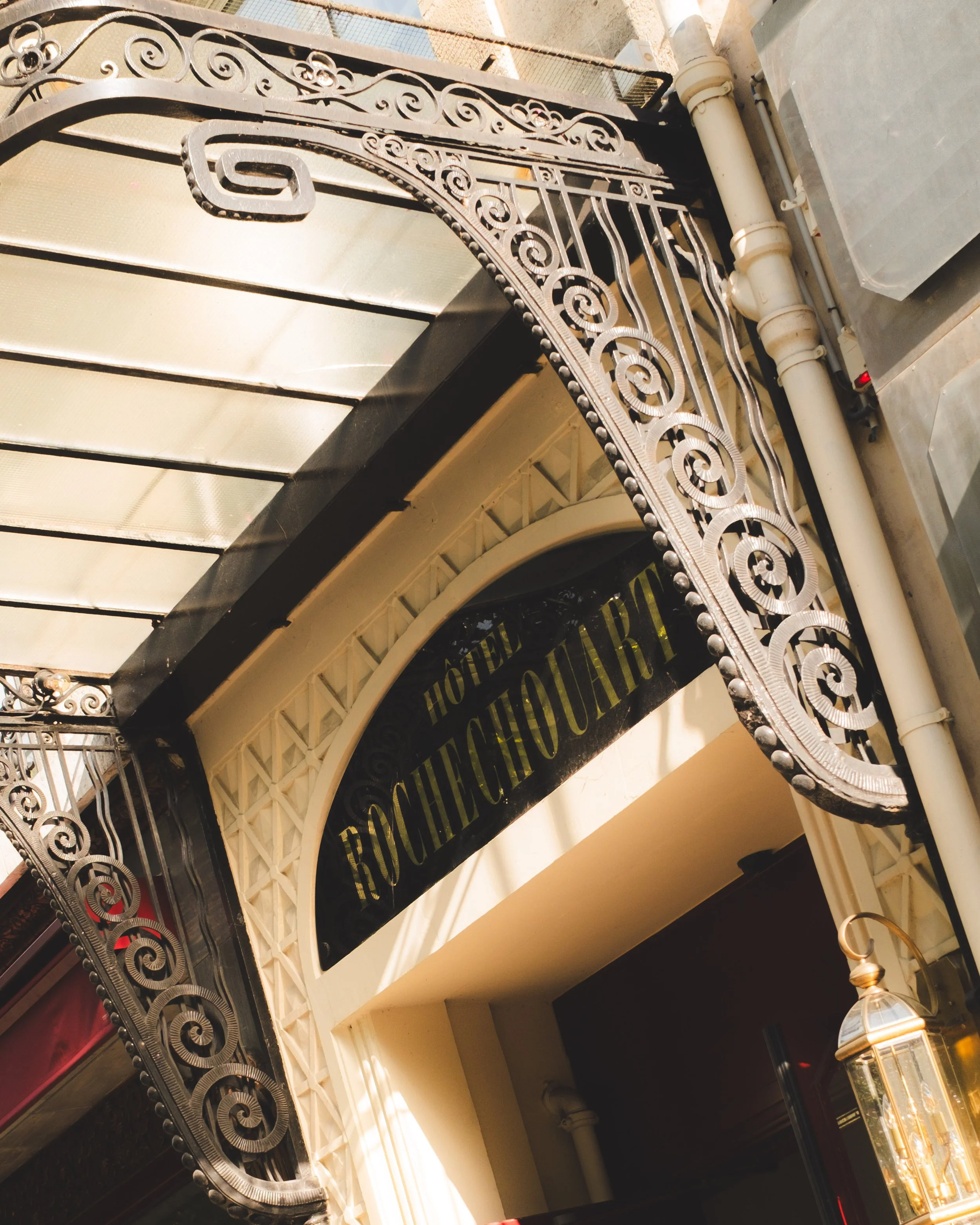 Close-up view of a black and gold sign with the words 'HOTEL ROCHECHOUART' partially visible, framed by ornate black wrought iron decorative elements, and a glass lantern hanging below.