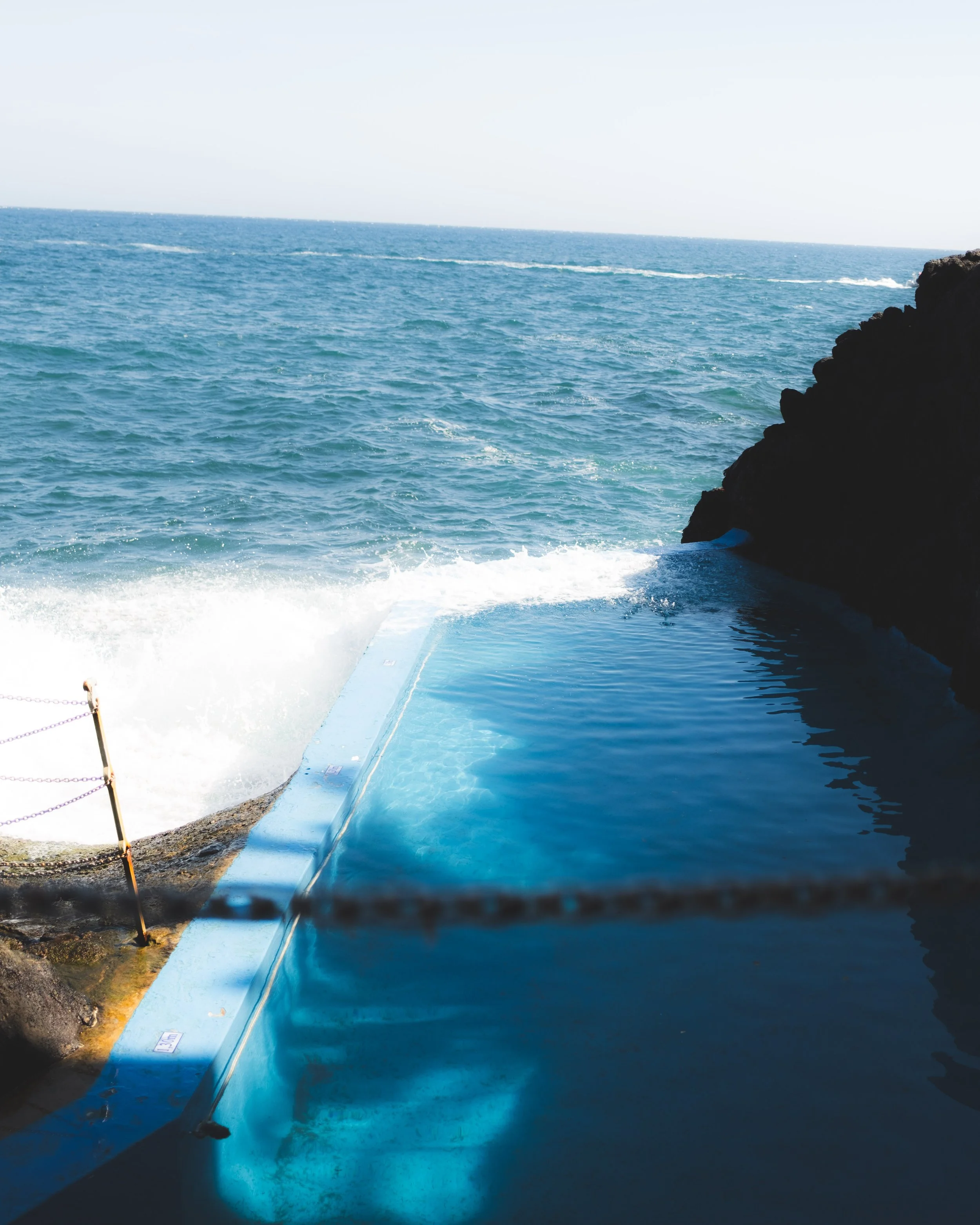 View of the ocean with waves hitting a rocky shoreline. A blue boat or vessel is partially visible in the foreground, with the water and horizon in the background.