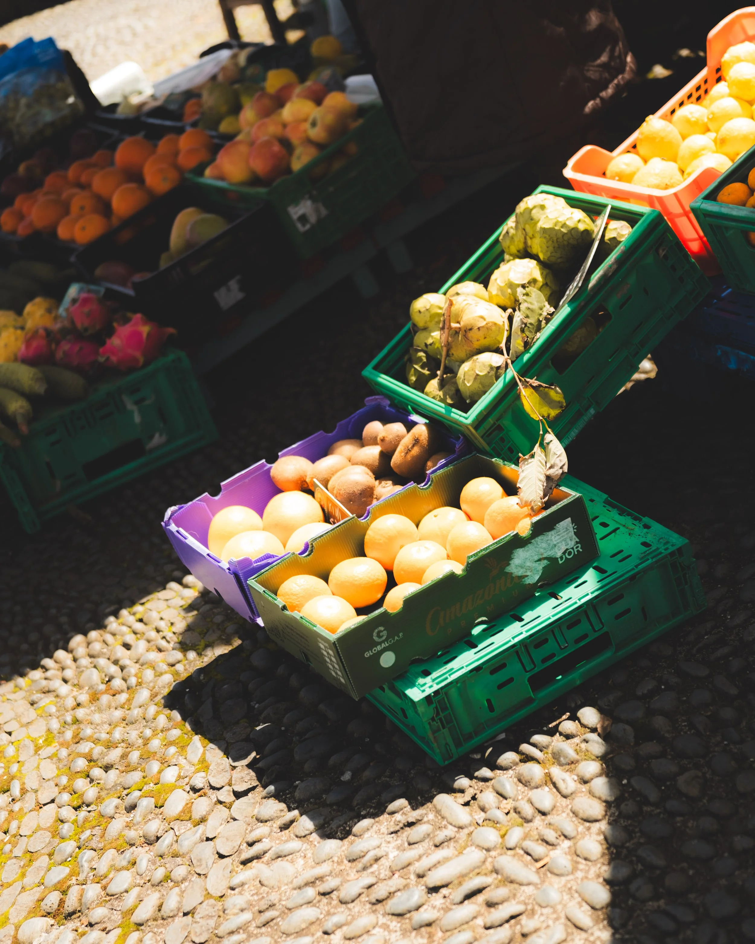 Arranged crates of fresh fruits including oranges, apples, kiwis, and possibly other seasonal produce at an outdoor market.
