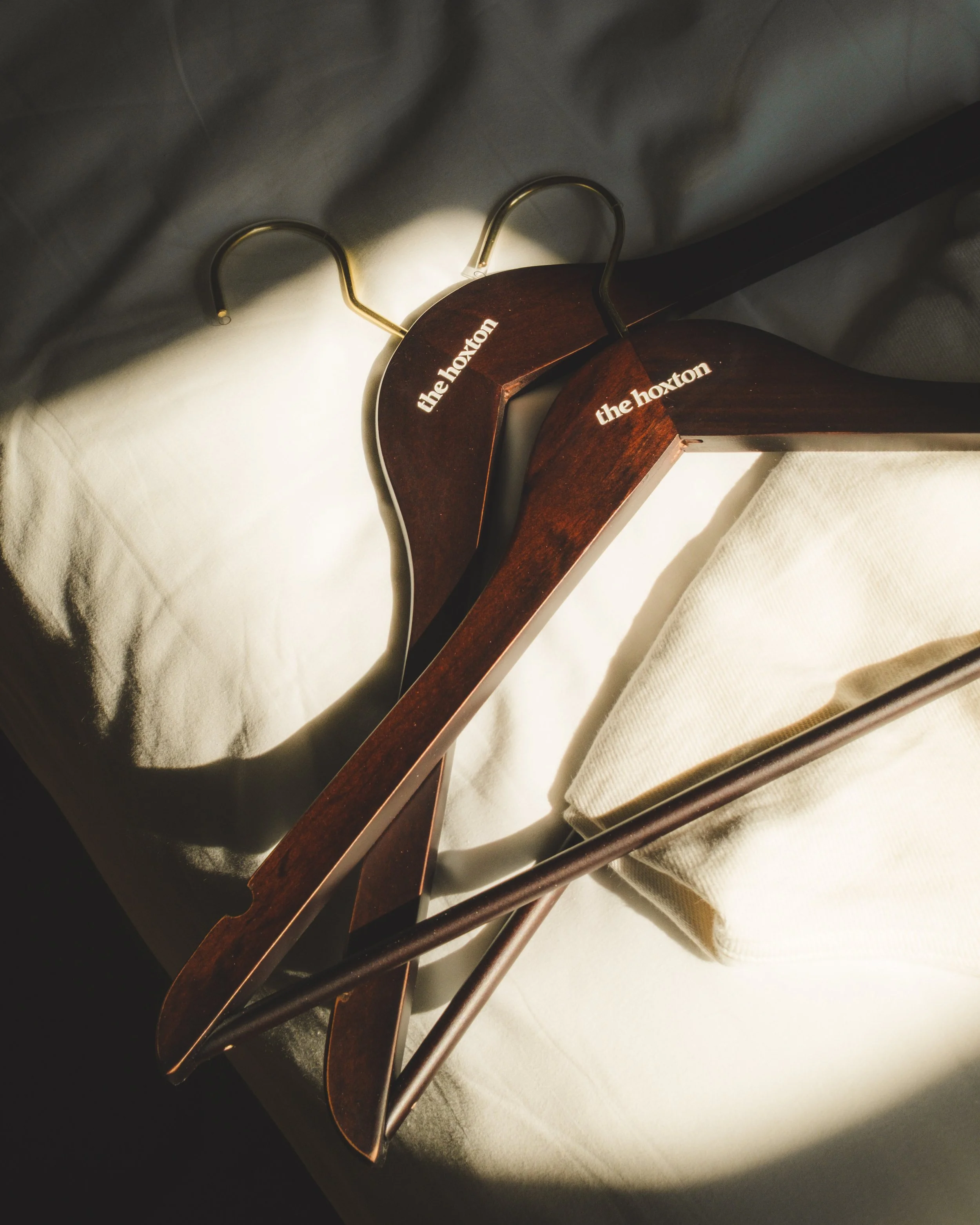 Two wooden hangers hanging on a white wall behind a gray fabric. The hangers have the words 'the hoxton' written on them in white.