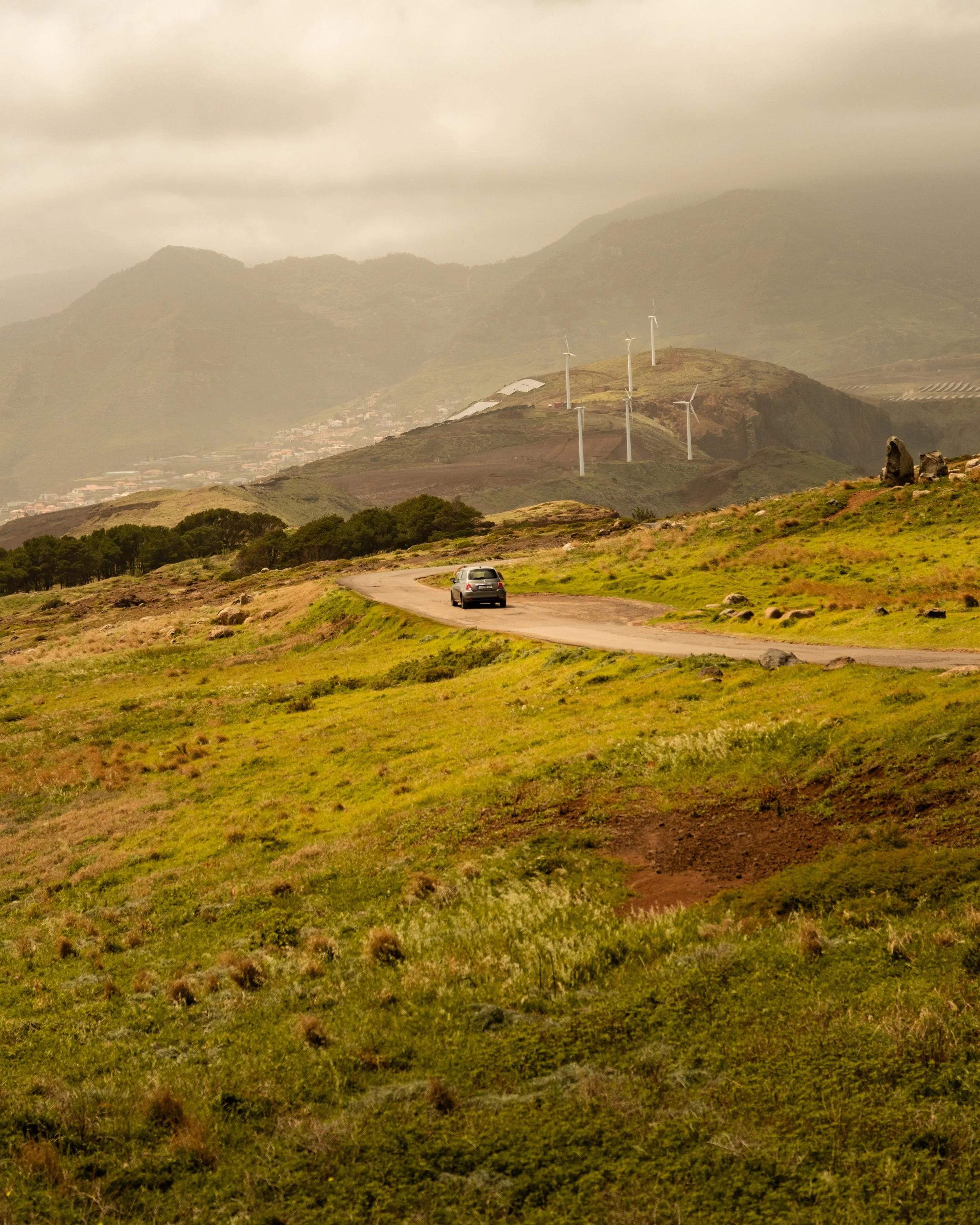 A rural landscape with green hills, a winding dirt road, wind turbines on a hill, and a car driving along the road. Mountainous terrain in the background under cloudy skies.