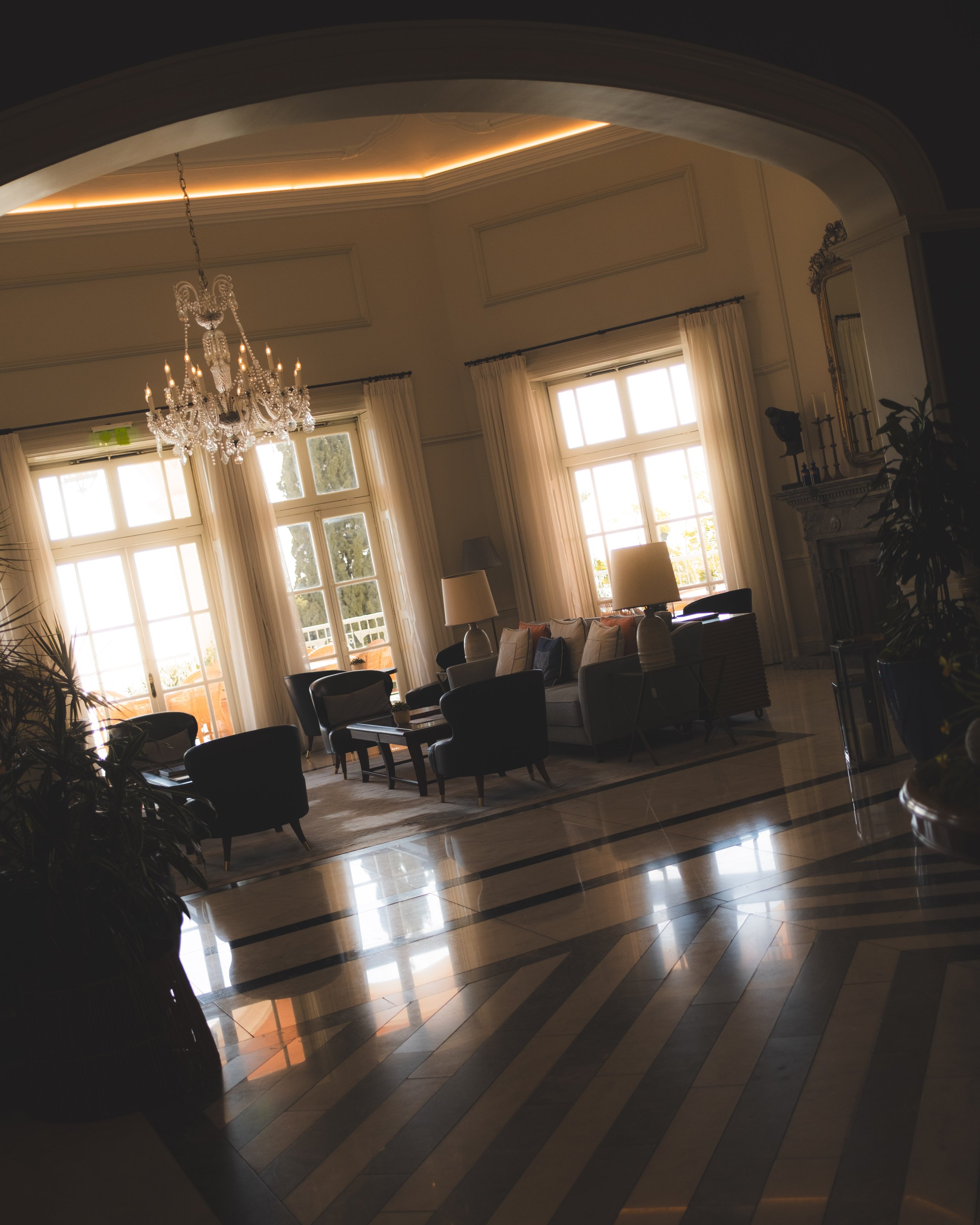 Elegant hotel lobby with large windows, white drapes, chandelier, beige walls, fireplace, and black chairs.