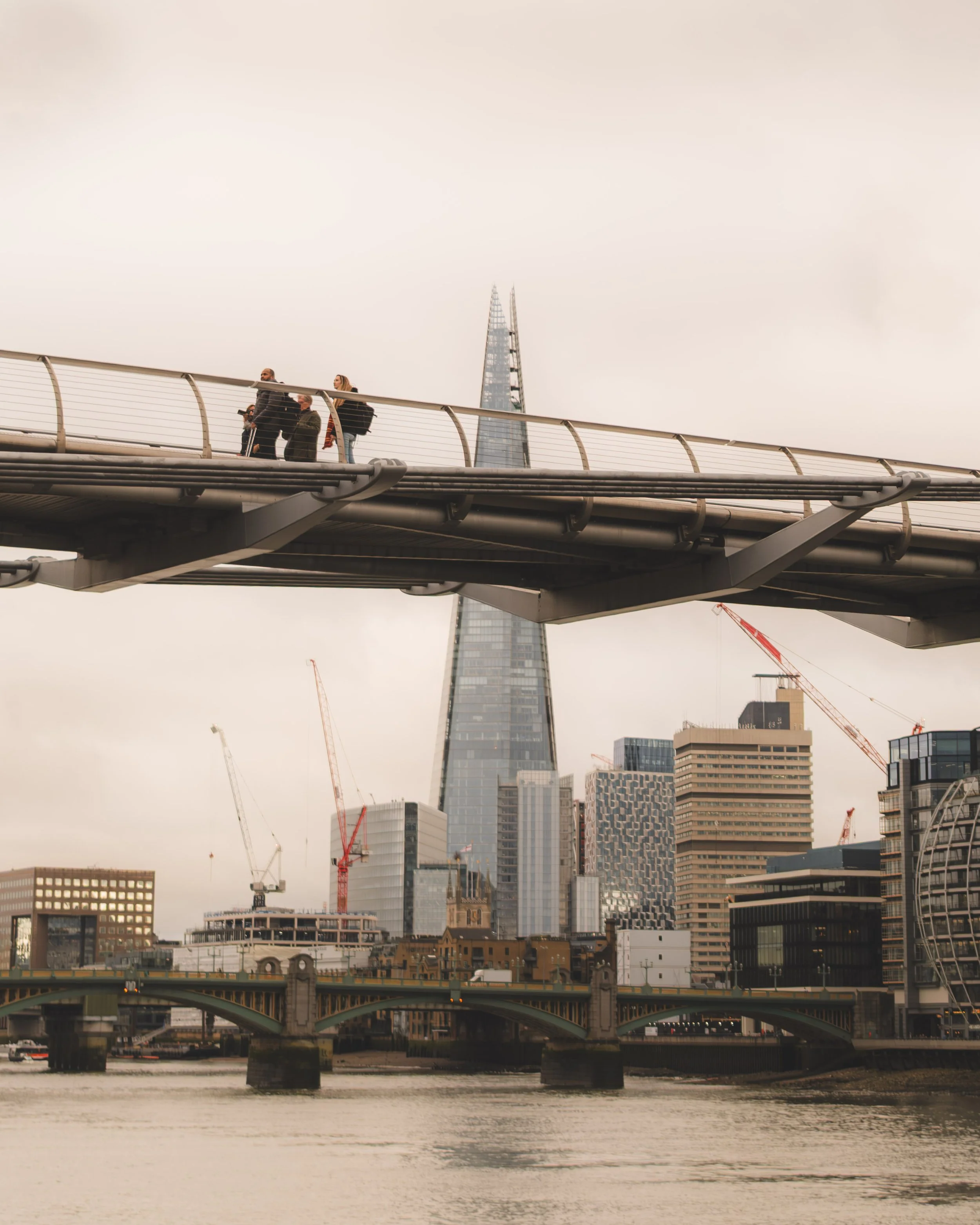 City skyline with modern skyscrapers, older buildings, a bridge over a river, and construction cranes, under a cloudy sky.