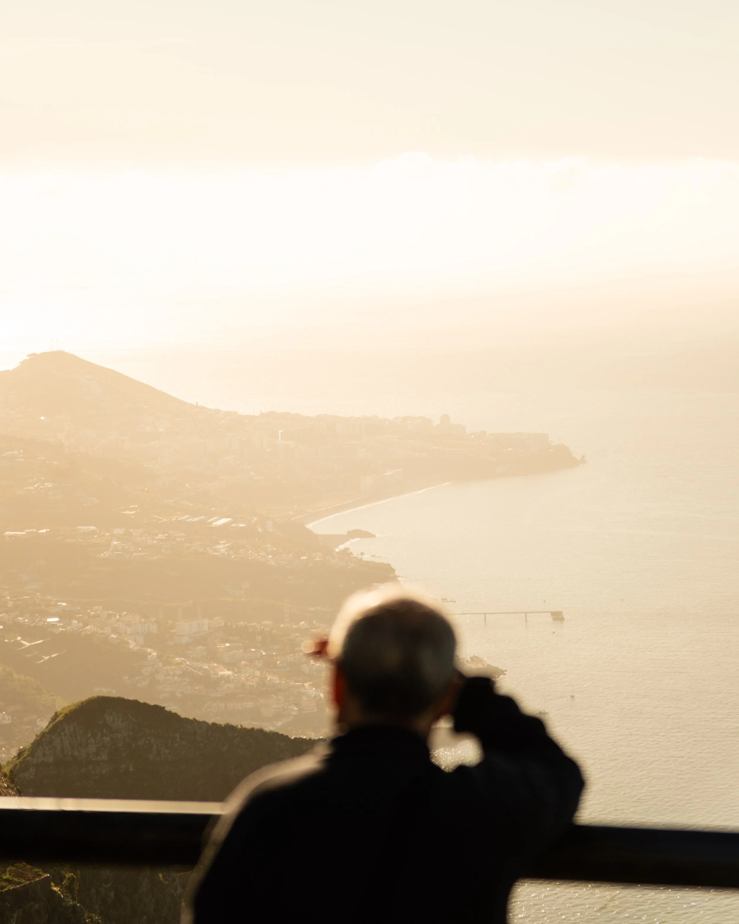 A person is sitting on a ledge facing a scenic view of hills and a city, with a hazy sunset or sunrise illuminating the landscape.