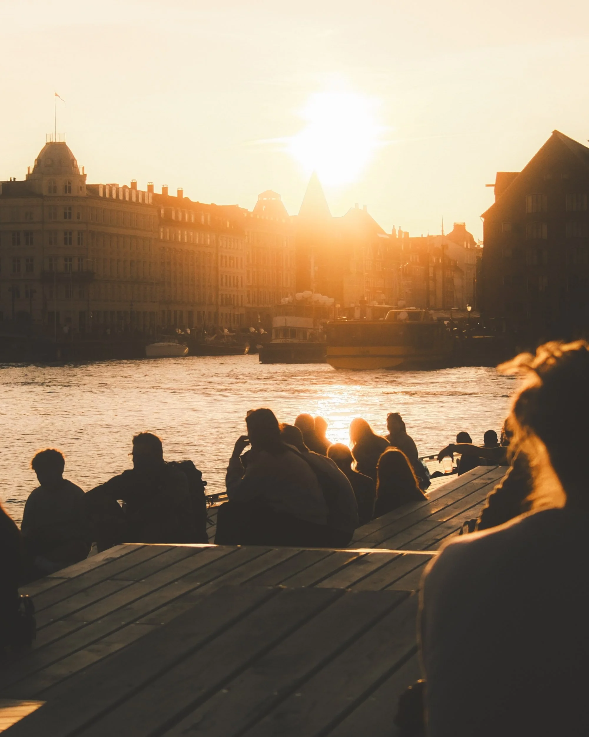 People sitting along a riverbank at sunset with city buildings and boats in the background.