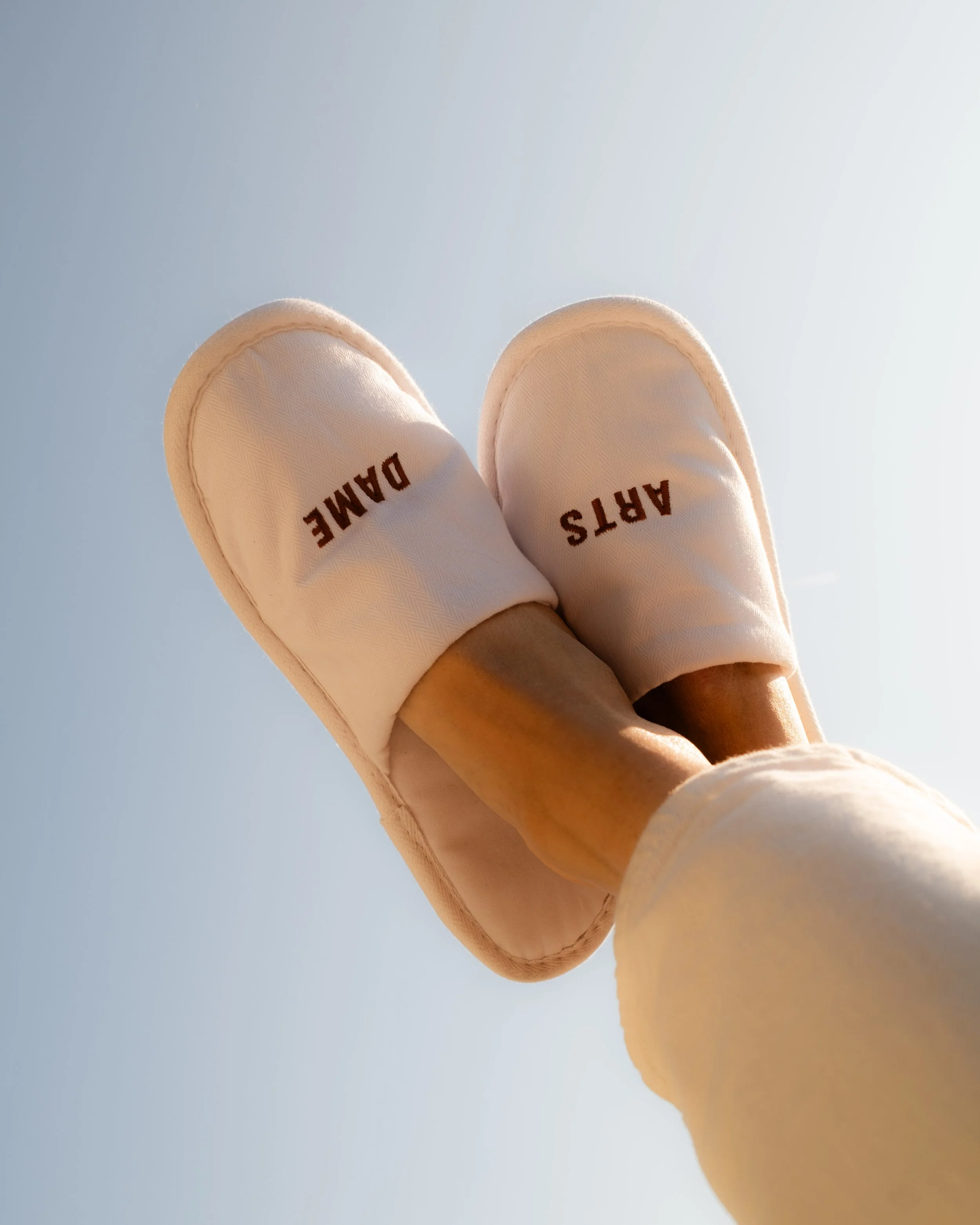 Close-up view of a person's feet wearing white slippers with the words "ARTS" and "DAME" written on them, against a blue sky background.