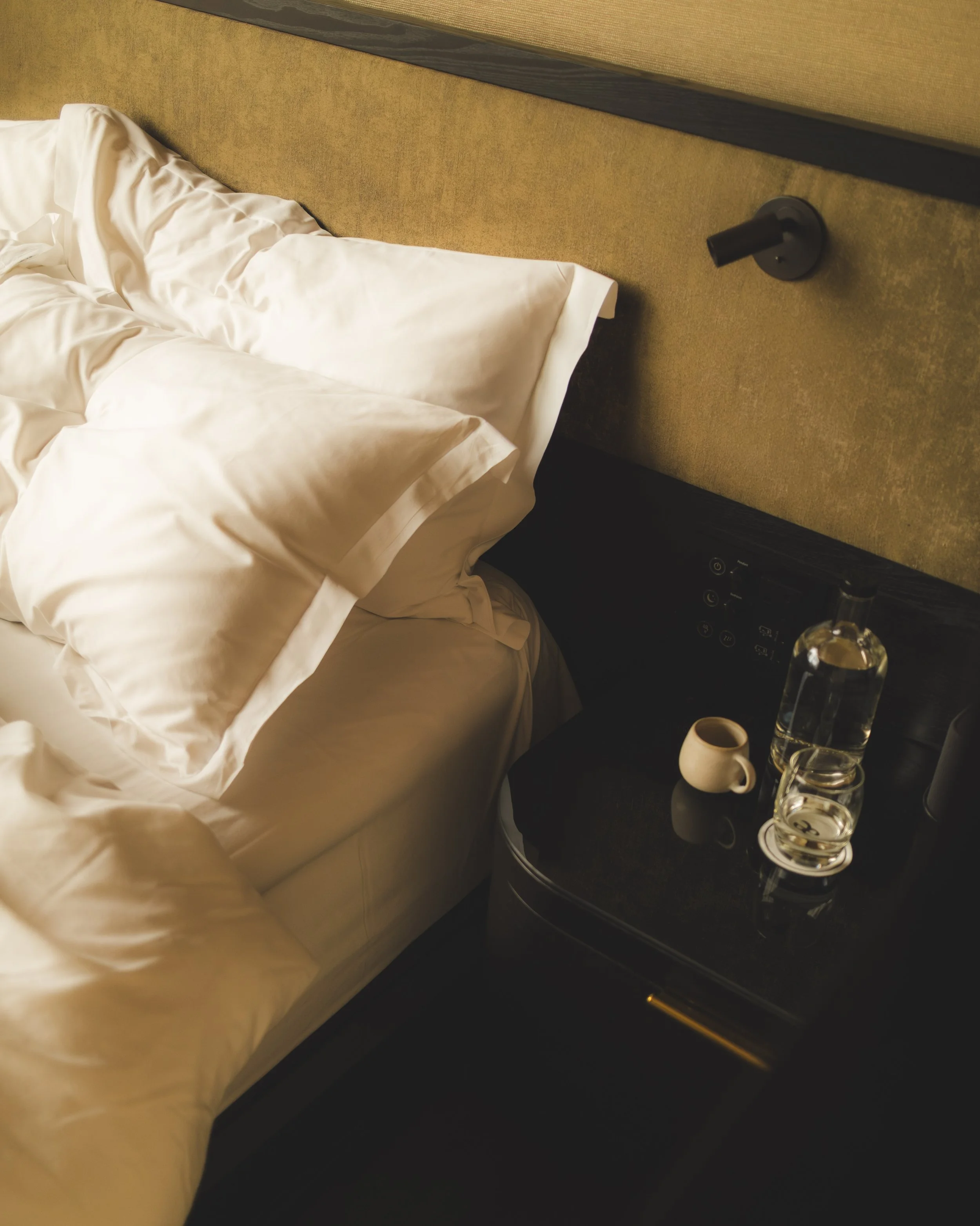 Close-up of a bed with white rumpled sheets and pillows, against a beige headboard. A black nightstand next to it holds a clear bottle of water, a white coffee mug, and a wine glass. Part of a control panel is visible at the bottom.