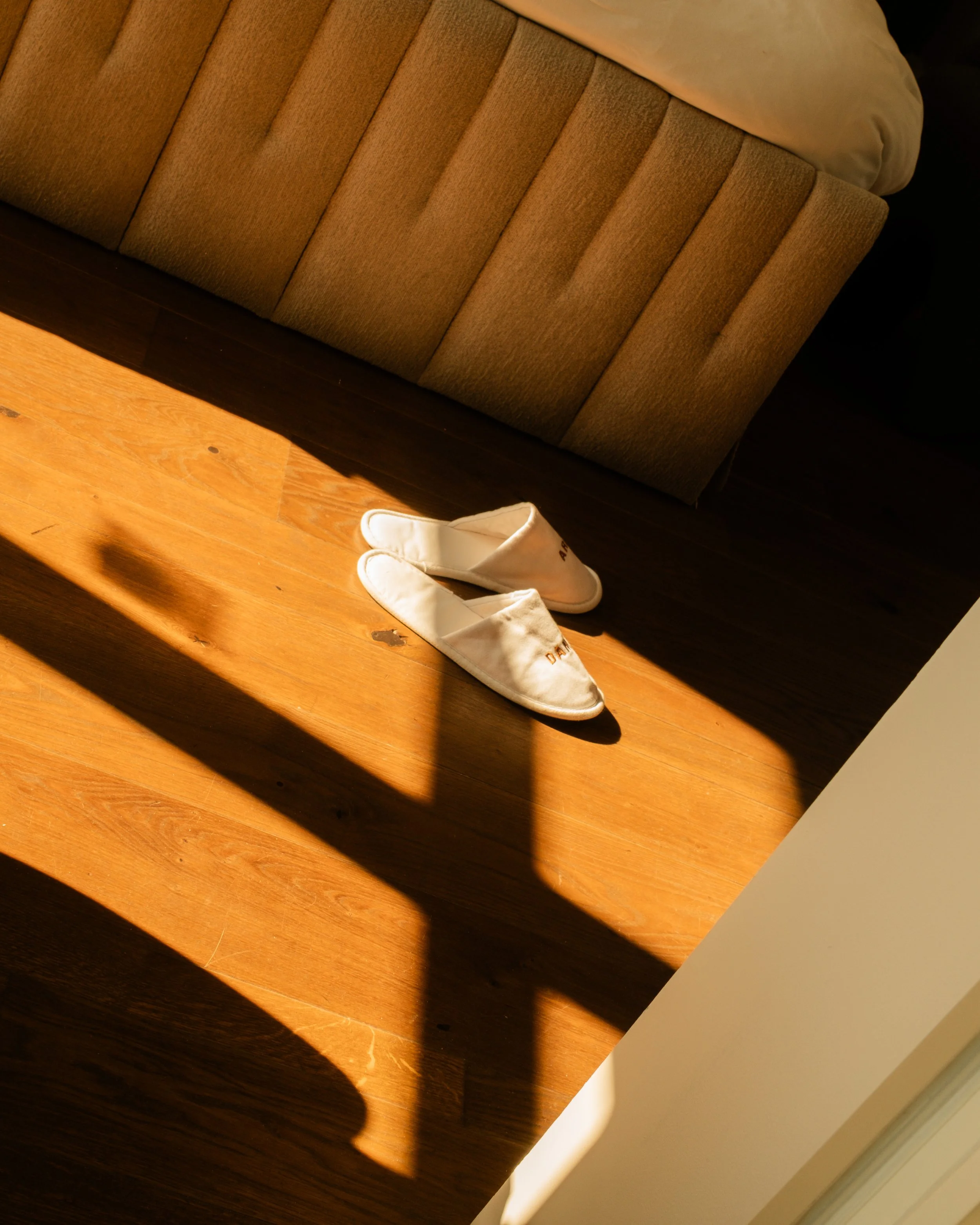 A pair of white slippers on a wooden floor near a bed with beige fabric upholstery. Lionel Chu - Hotel & Travel Photographer