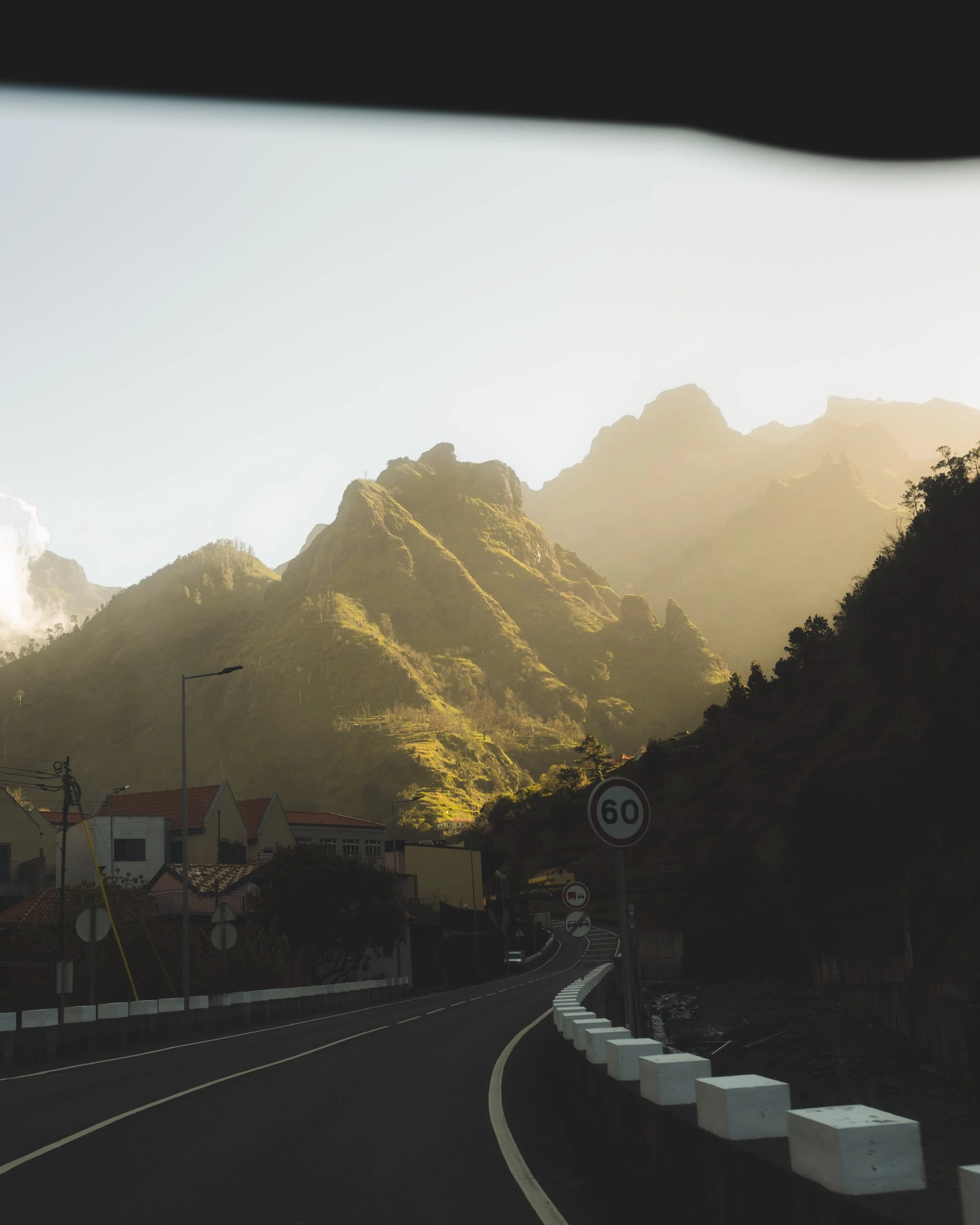A mountain landscape with a winding road in the foreground, residential houses with red-tiled roofs, street signs, and a mountain range in the background during sunset.