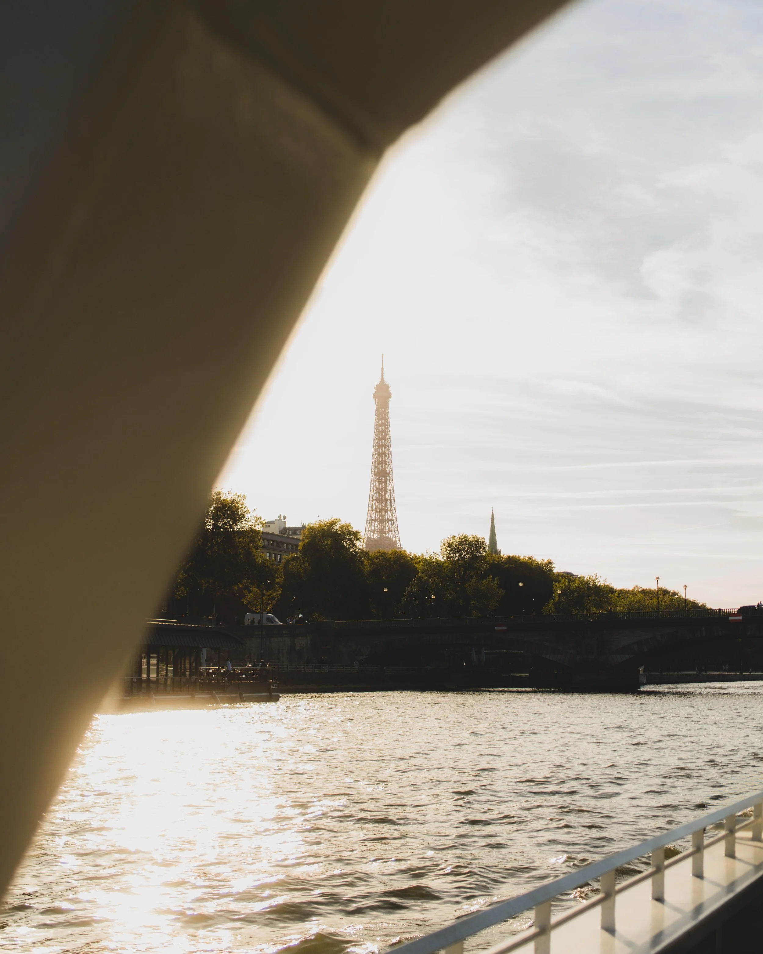 View of the Eiffel Tower through a boat window with water, trees, a bridge, and the Paris skyline in the background during sunset. Lionel Chu - Hotel & Travel Photographer