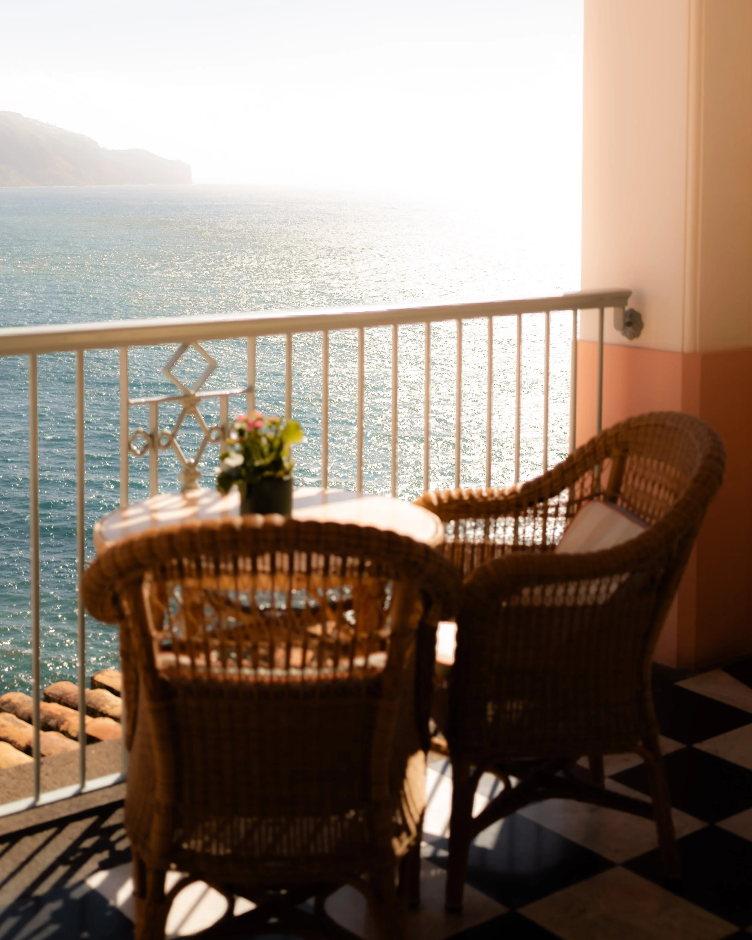 A balcony with wicker chairs and a small round table with a flowerpot, overlooking a sparkling body of water and distant hills or islands, bathed in bright sunlight.