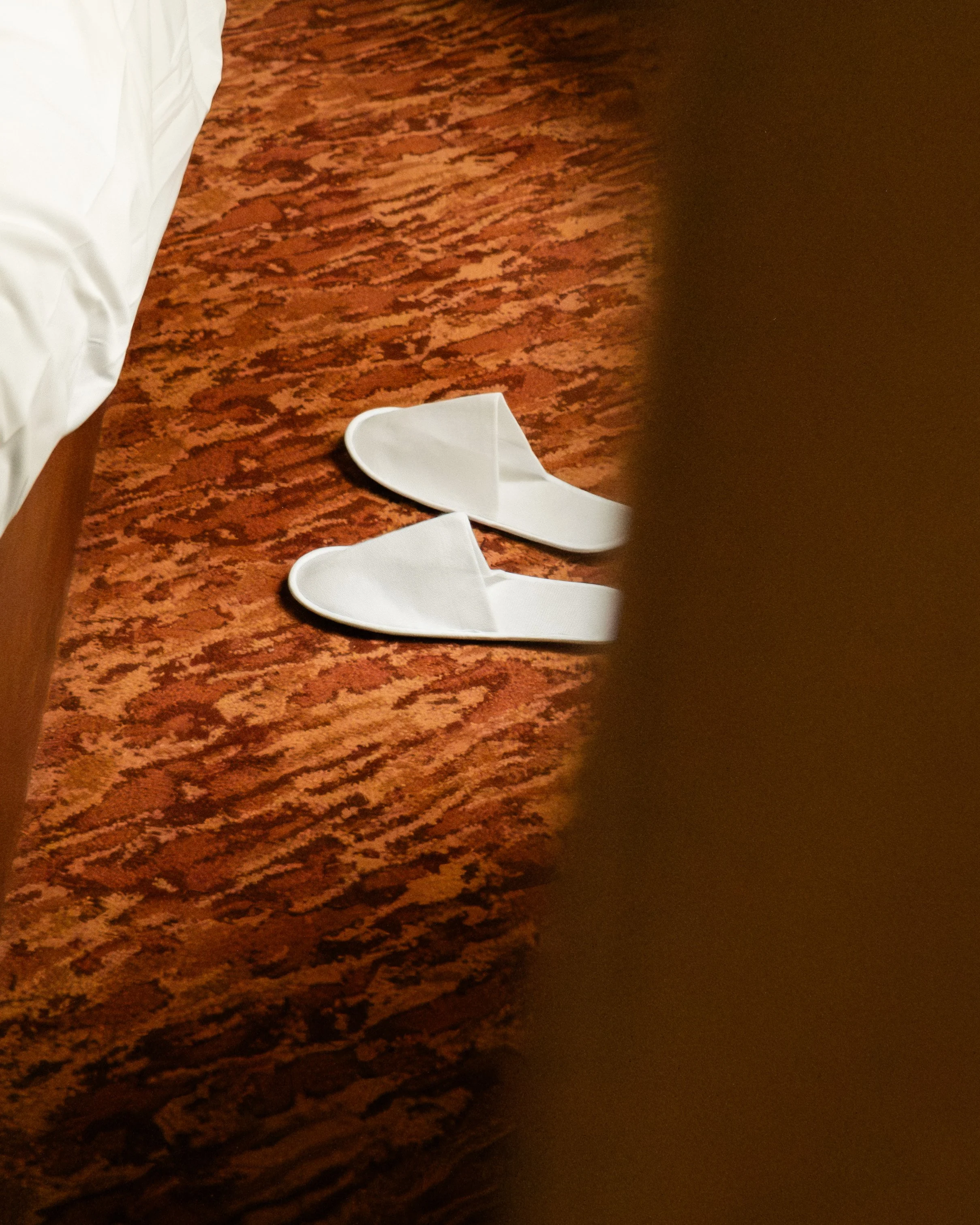 Pair of white slippers on a patterned red-brown carpet near the edge of a bed with white bedding. Lionel Chu - Hotel & Travel Photography