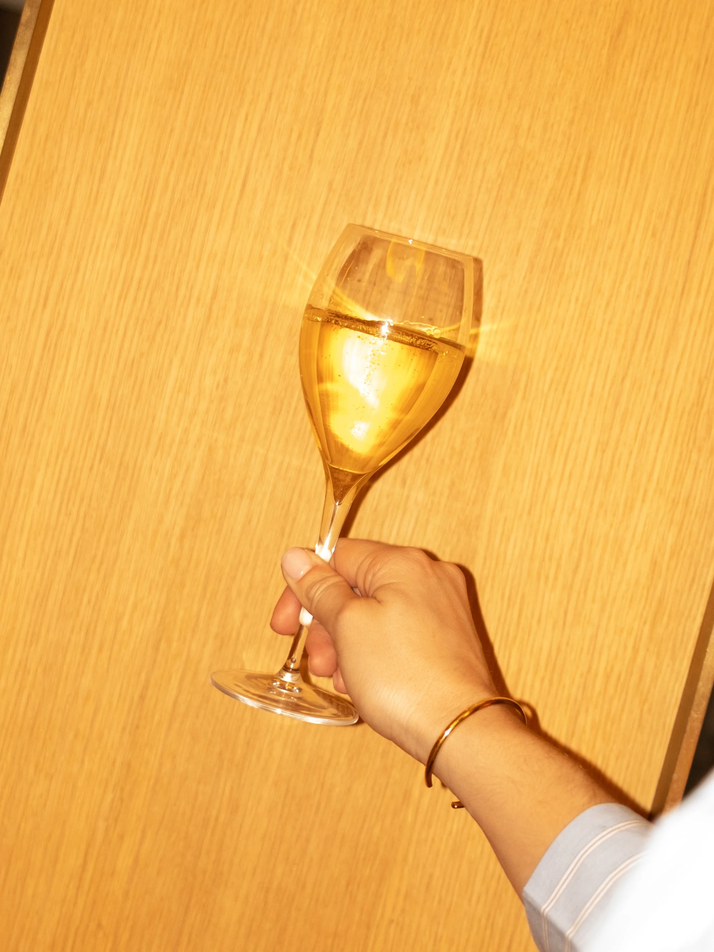 Hand holding a champagne flute filled with sparkling white wine against a wooden background. Lionel Chu - Hotel & Travel Photography