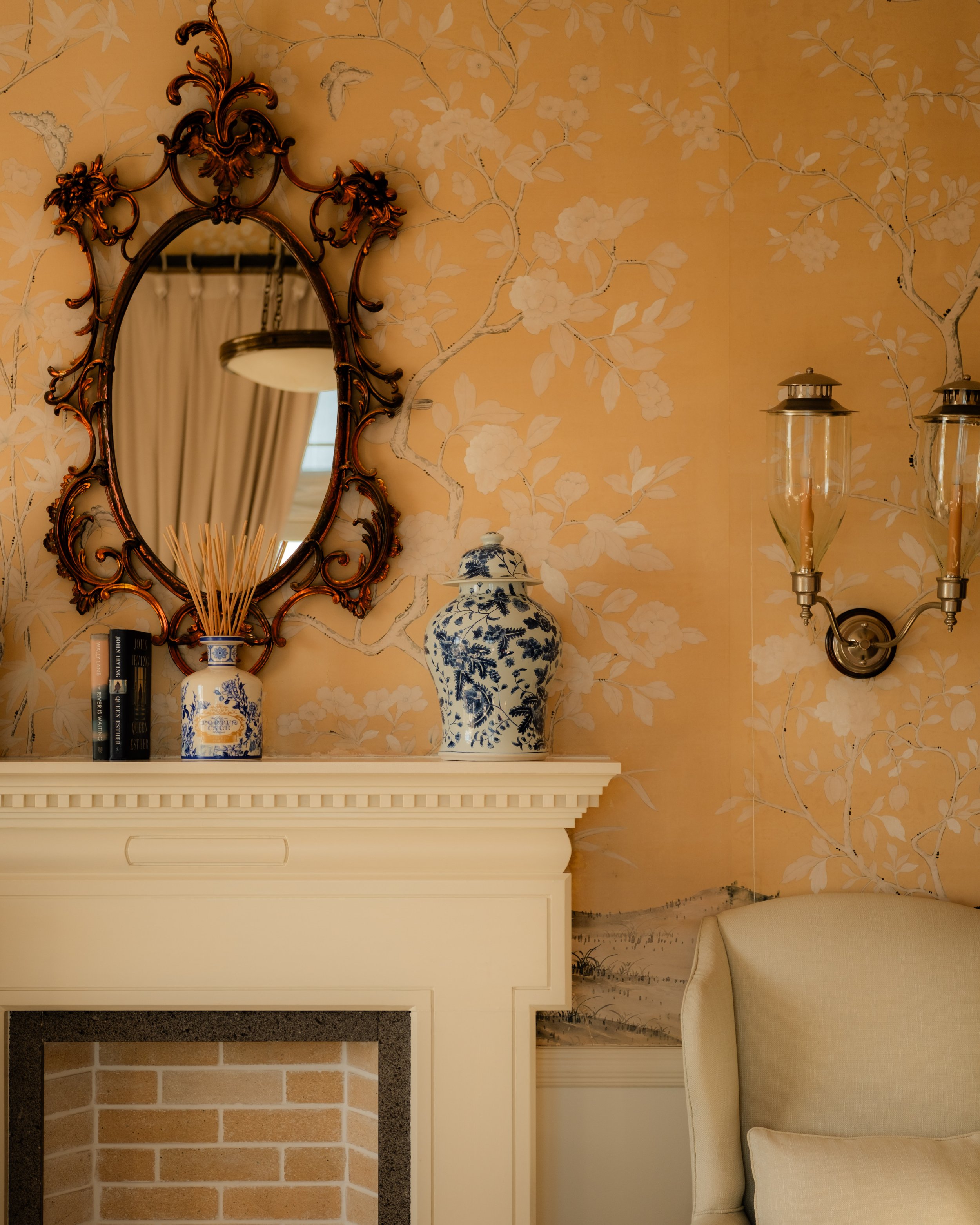 Living room with yellow floral wallpaper, an ornate gold-framed mirror, blue and white Chinese porcelain vases, a white fireplace mantel with books, and a cream-colored sofa.