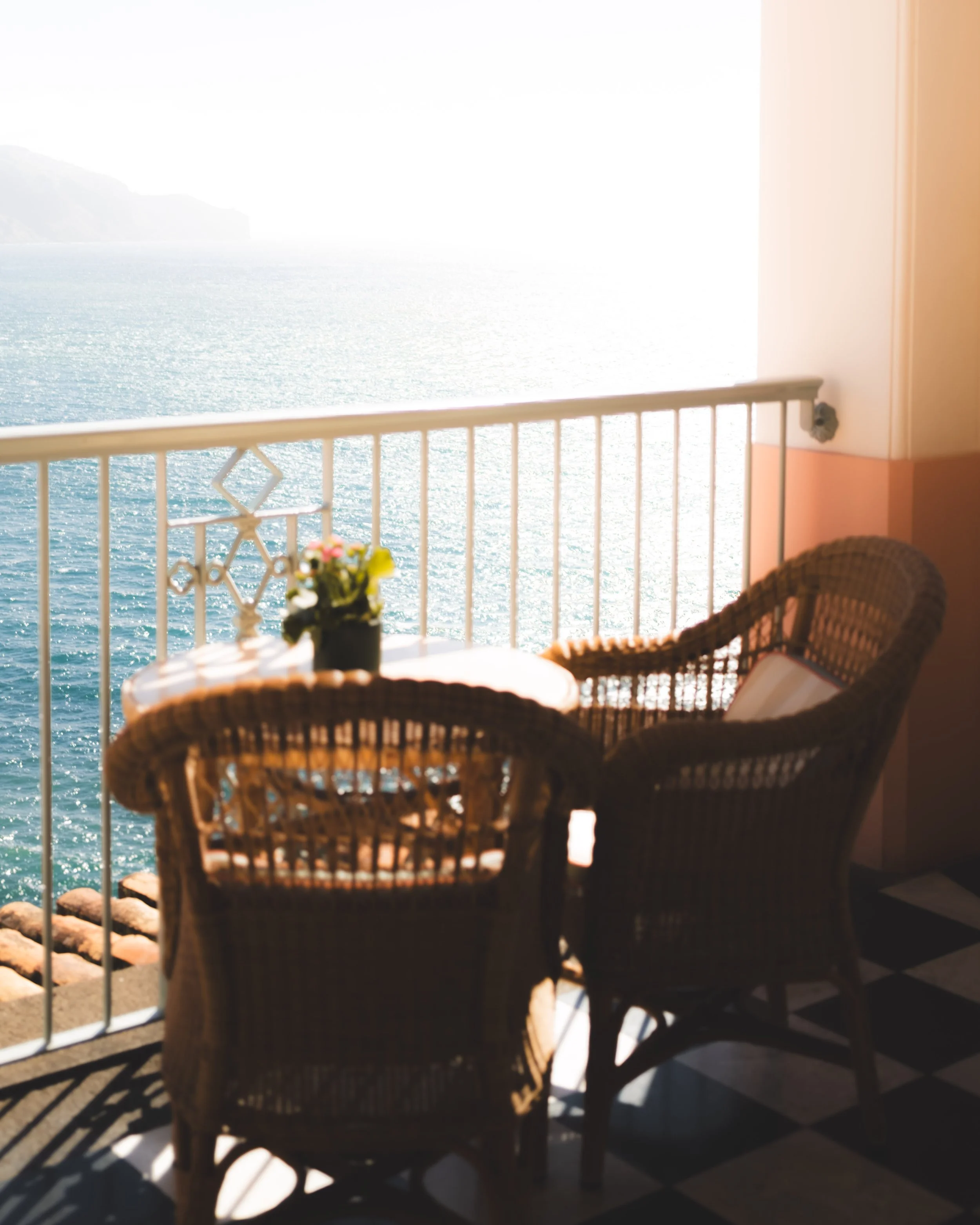 Two wicker chairs and a table with a small potted plant with pink, yellow, and white flowers on a balcony overlooking the ocean, with distant land visible on the horizon.