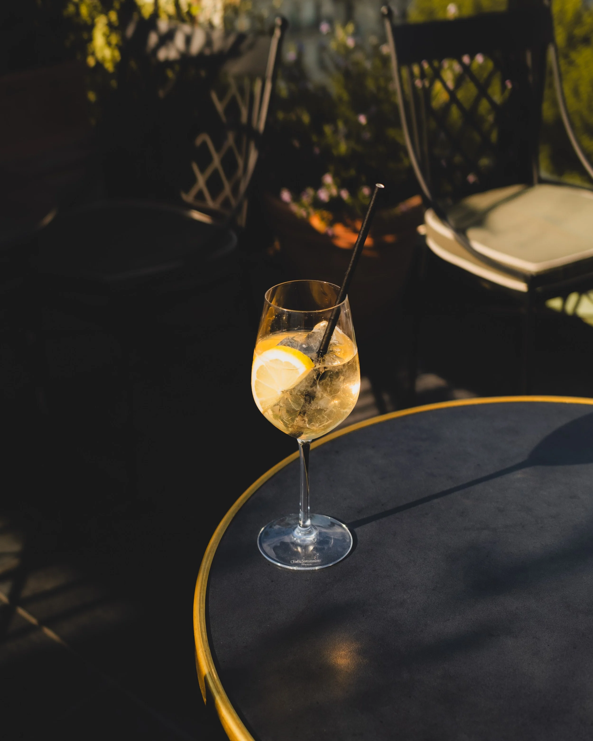 A cocktail glass with lemon slices and ice, a straw, placed on a dark surface outside, with blurred green foliage and sunlight in the background.