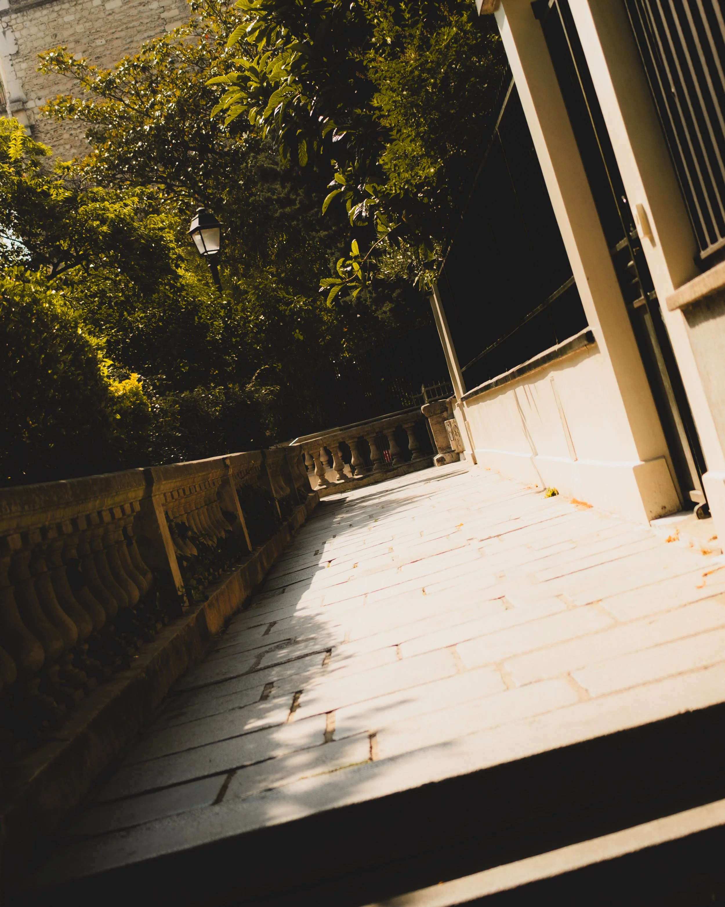 Photograph of a stone-paved sidewalk lined with a decorative stone railing, surrounded by green bushes and trees, with a black metal fence on the right and a streetlamp visible in the distance, taken during daylight. Lionel Chu - Hotel & Travel Photo