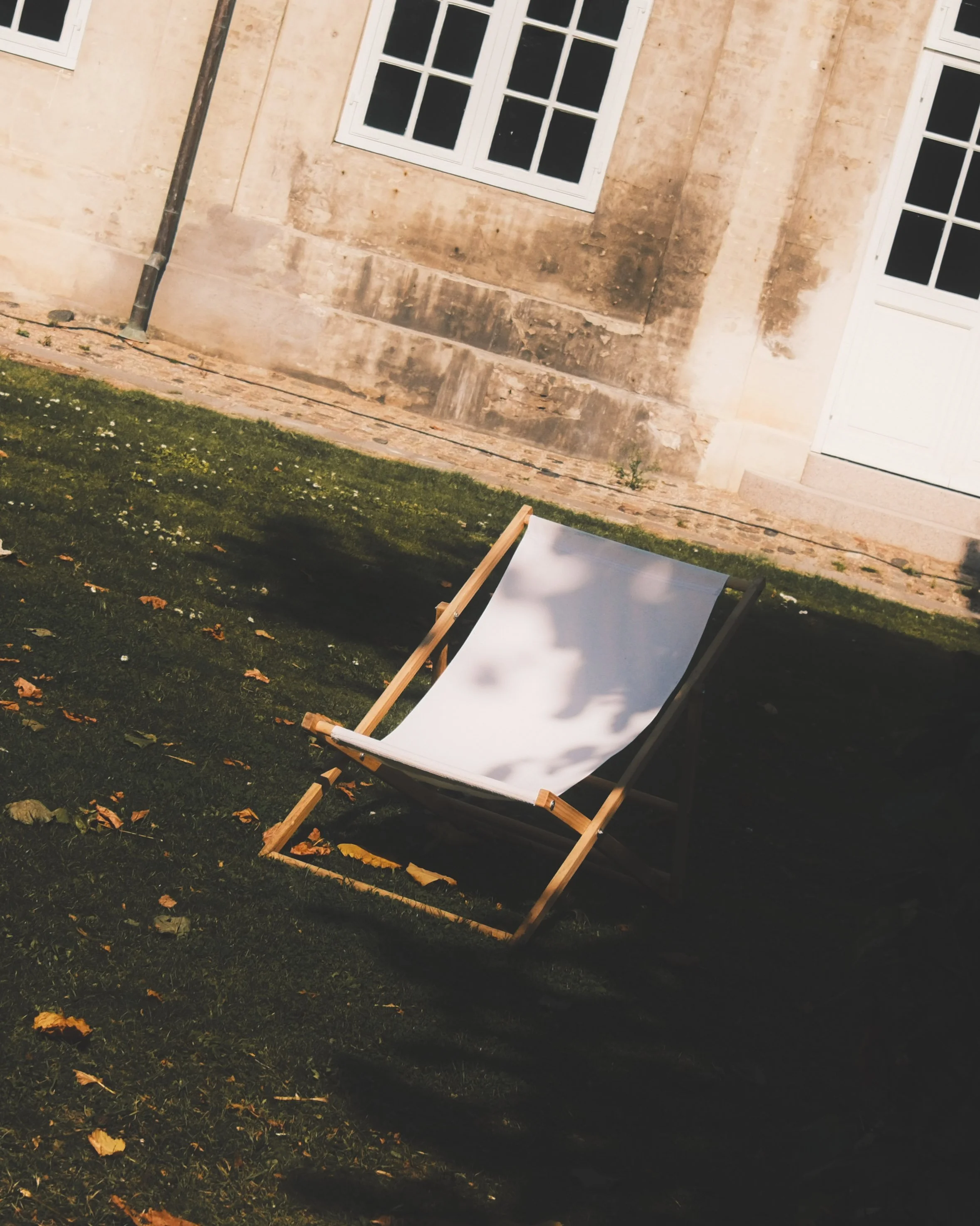 Daytime outdoor scene featuring a white folding chair on grass, fallen leaves, an old stone building with a large window, and a metal pole.