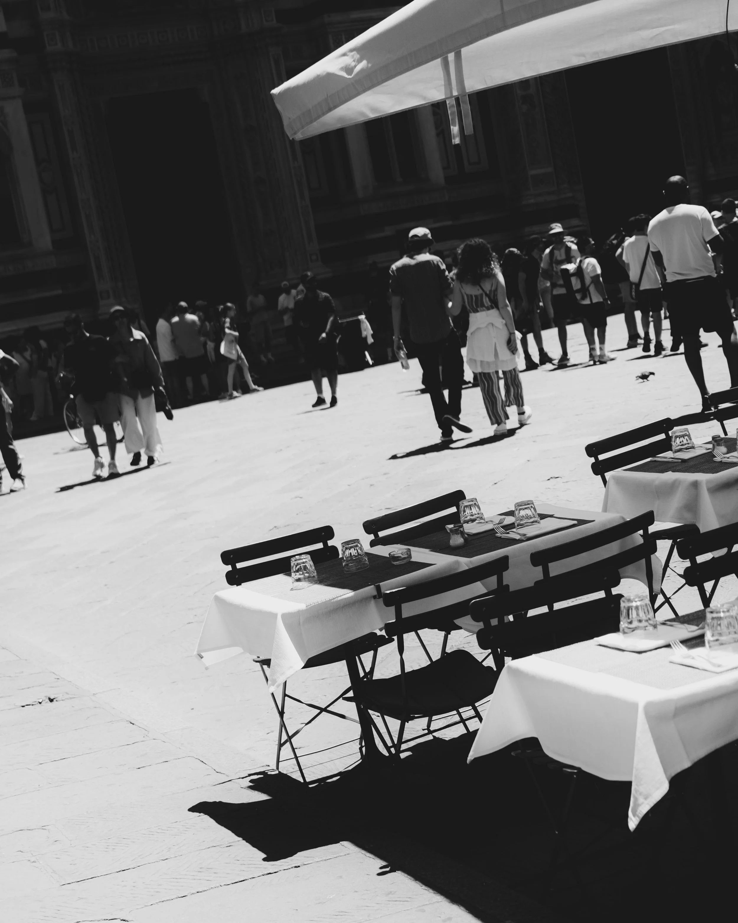 Outdoor terrace with tables and chairs, set with tablecloths, glasses, and utensils, overlooking a busy plaza with pedestrians and historic architecture. Lionel Chu - Hotel & Travel Photographer
