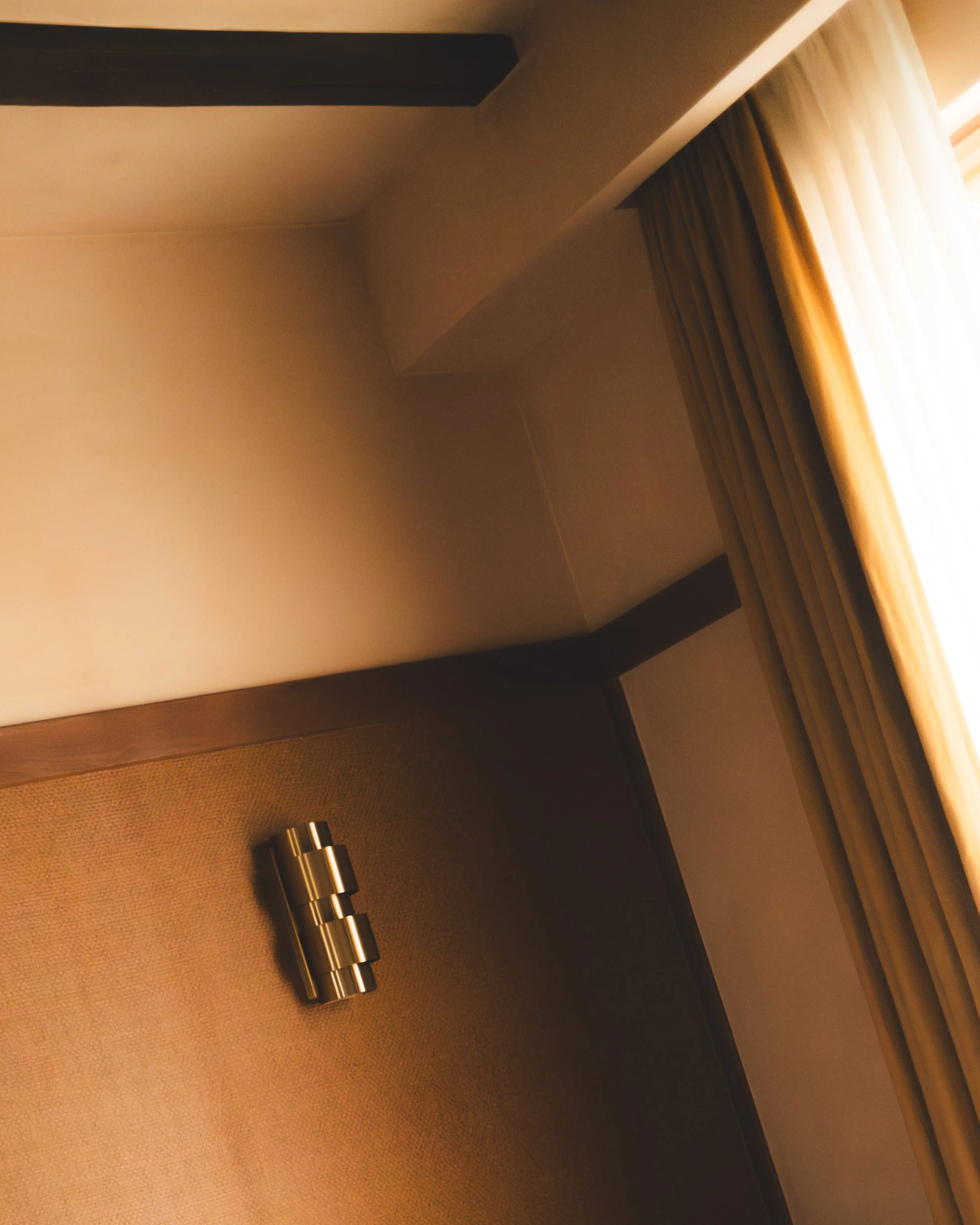 Hotel room desk area with a metallic cylindrical lamp on the dark wooden table, window with curtains, and textured wall.