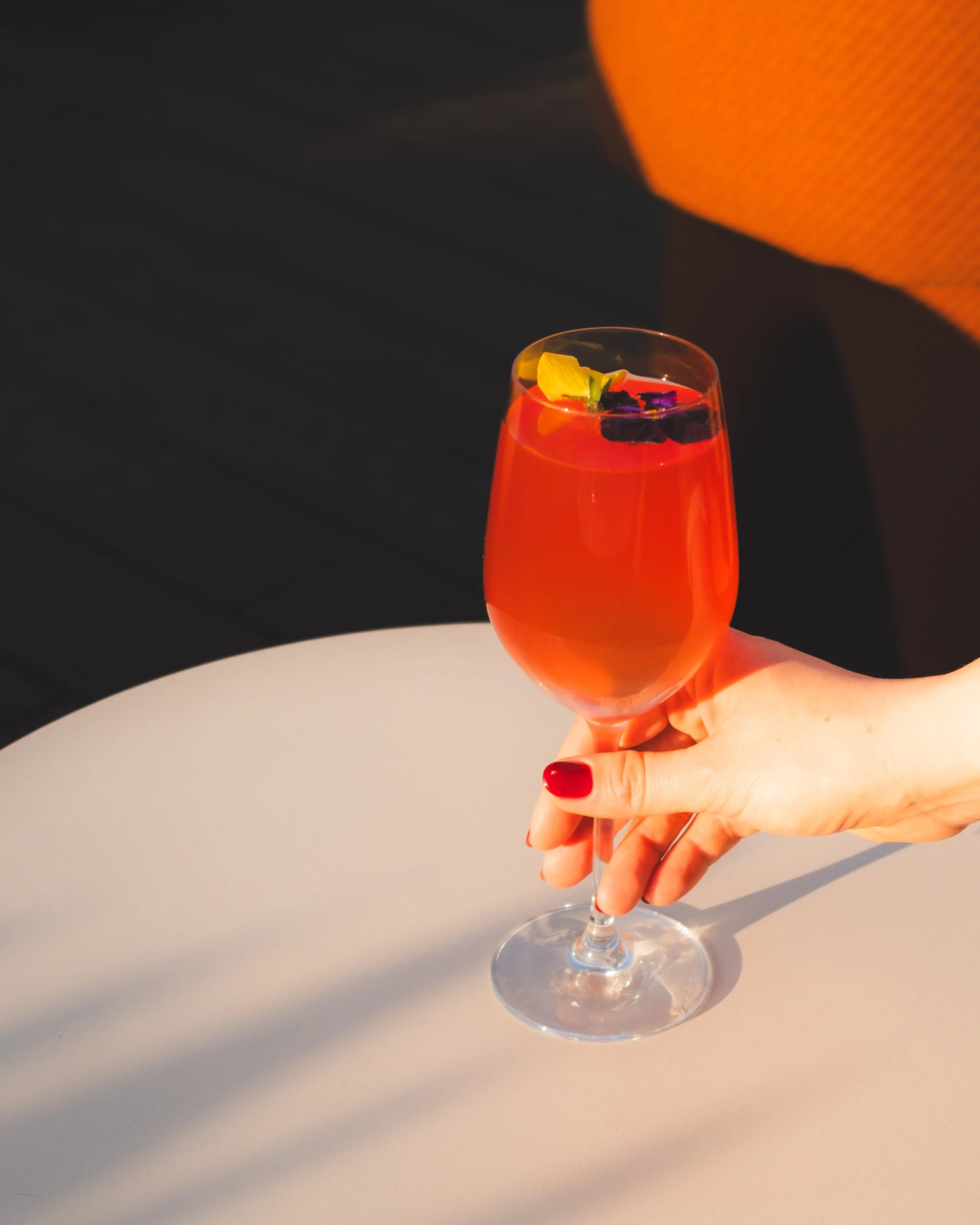 A close-up of a hand holding a cocktail glass with a red-orange drink, garnished with a lemon wedge and purple edible flowers, against a dark background with a white surface.