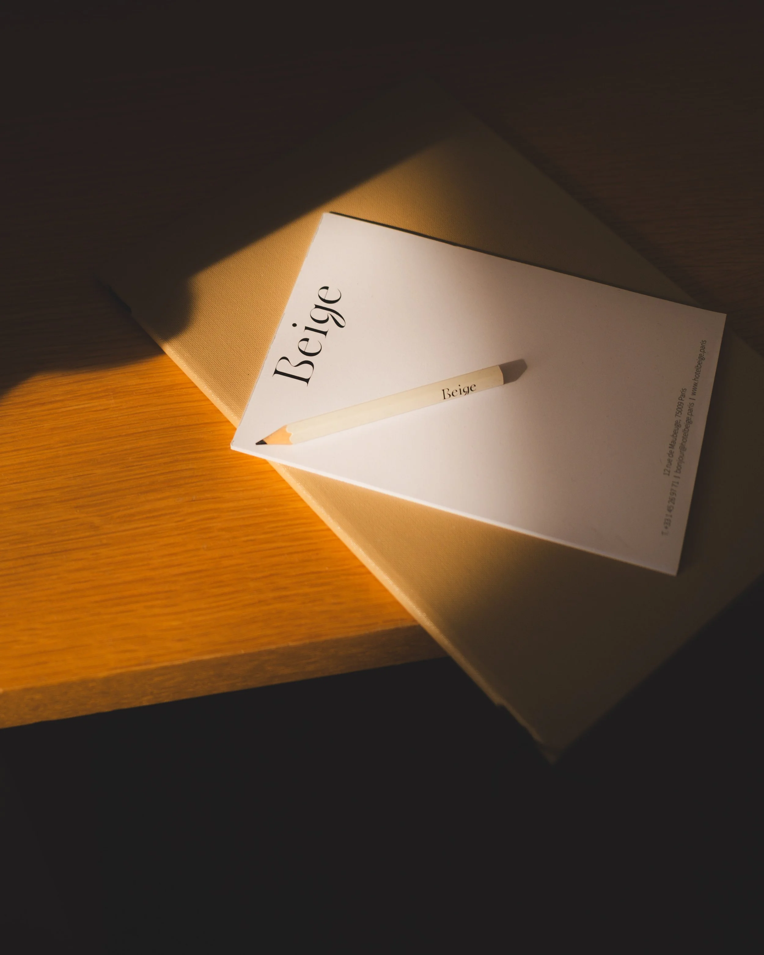 A book titled 'Beige' resting on a beige fabric-covered surface, with a beige-colored pencil nearby, both on a wooden table under warm lighting.