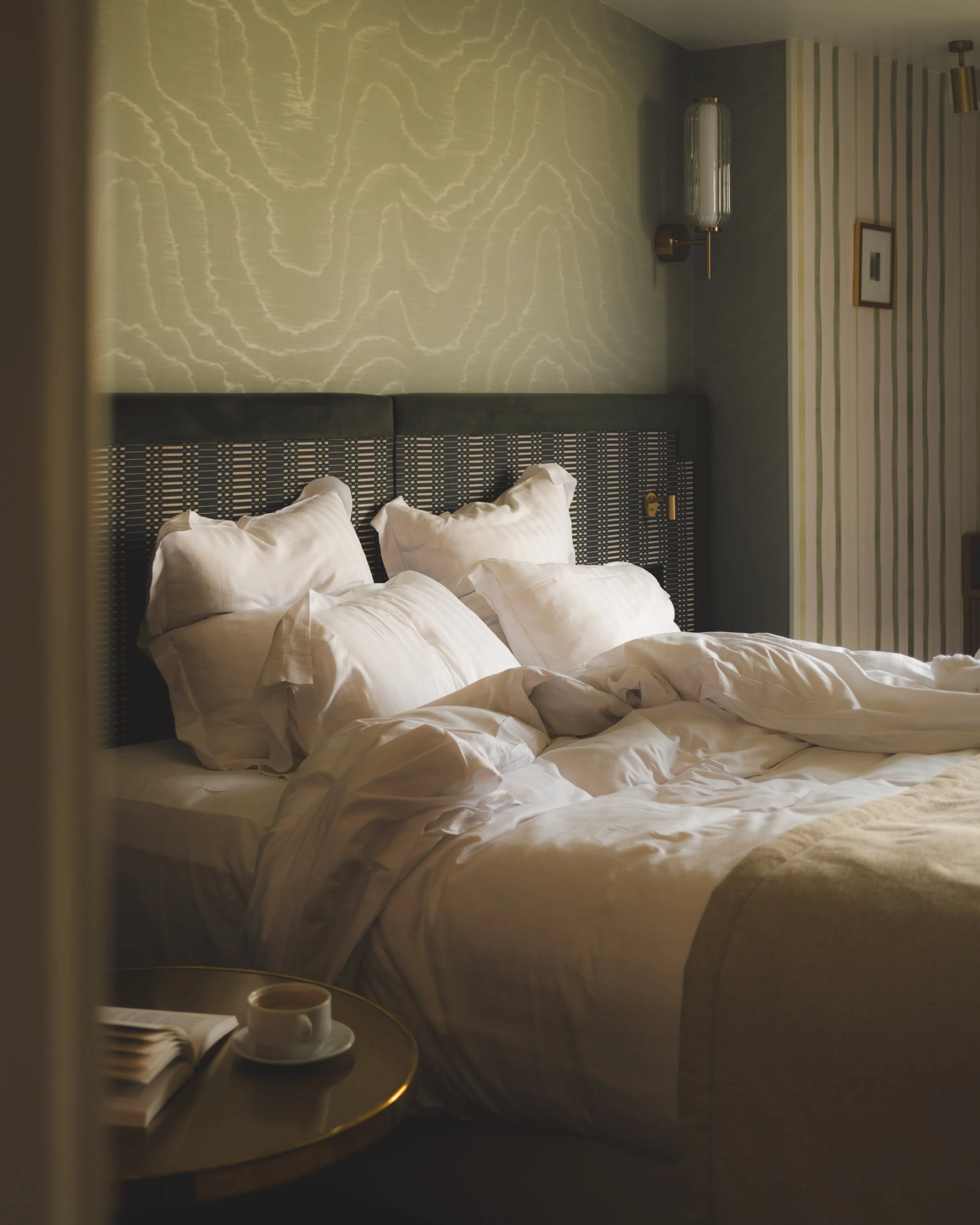 Hotel room with bed, pillows, and a side table with a cup of coffee and an open book.