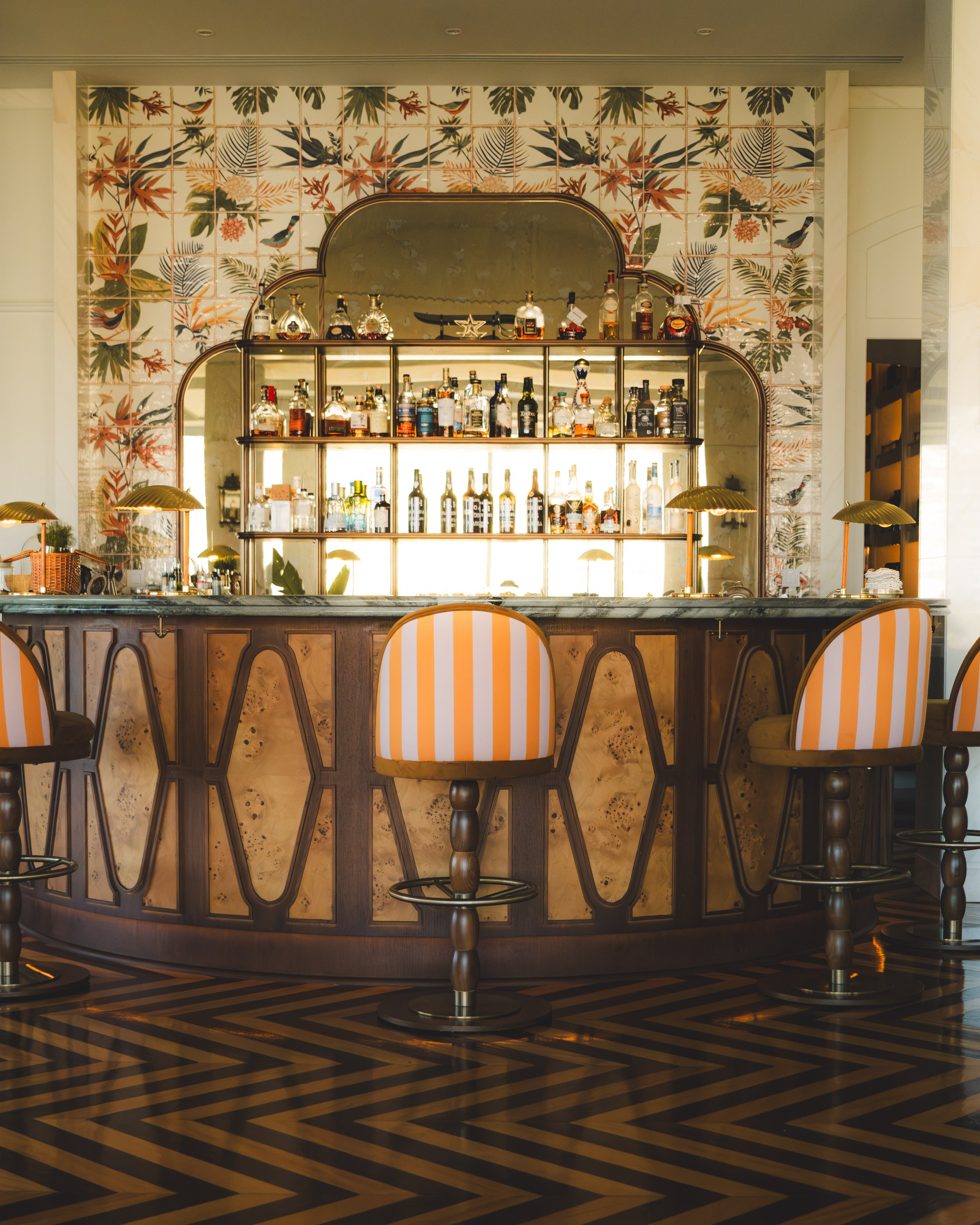 A bar with a curved wooden counter, bar stools with striped upholstery, a backlit shelf of various liquor bottles, a decorative mirror, and a tropical-themed tiled wall in the background.