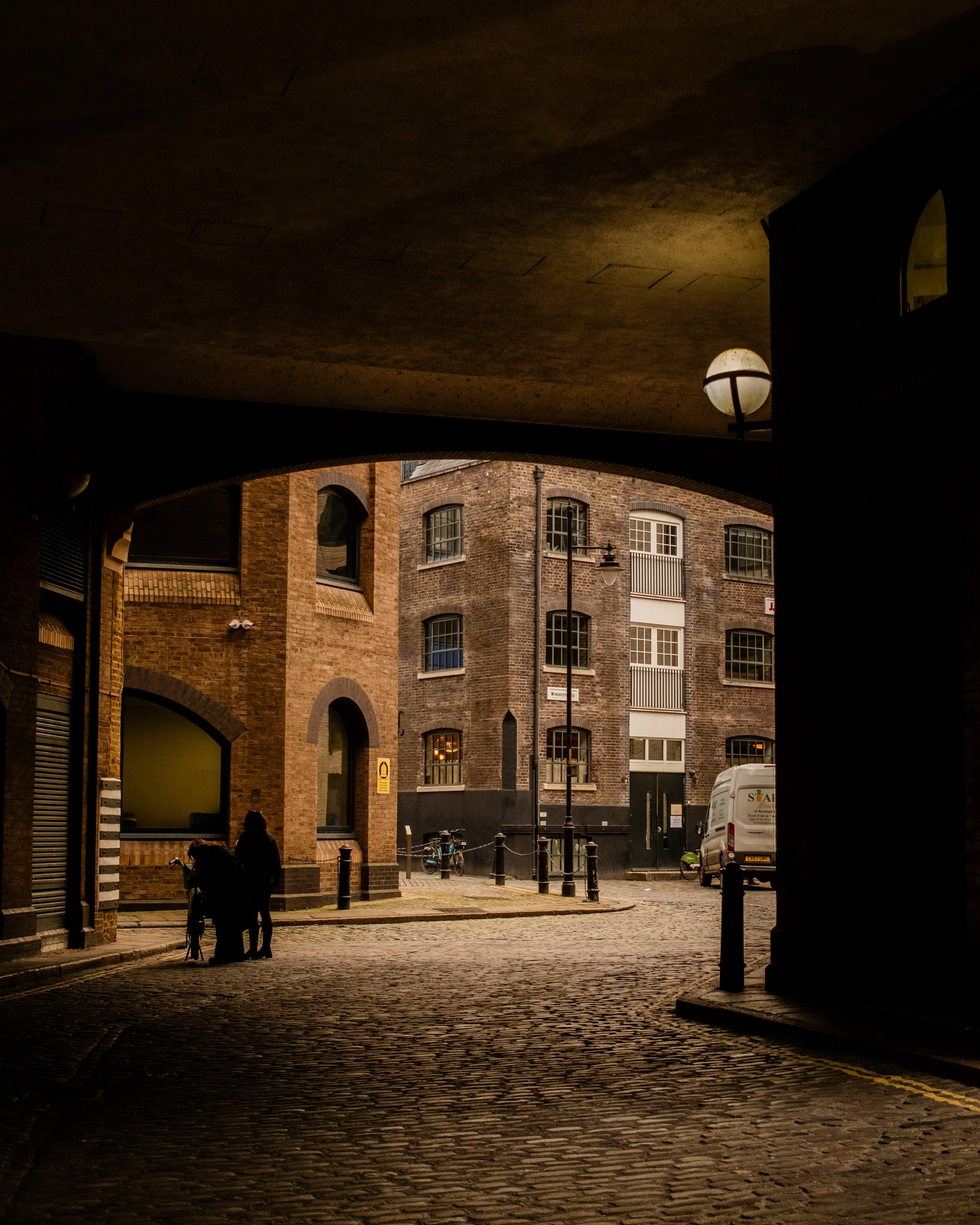 View of a cobblestone street under a bridge with brick apartment buildings in the background, a man and woman with a stroller and a bicycle parked on the sidewalk. Lionel Chu - Hotel & Travel Photographer