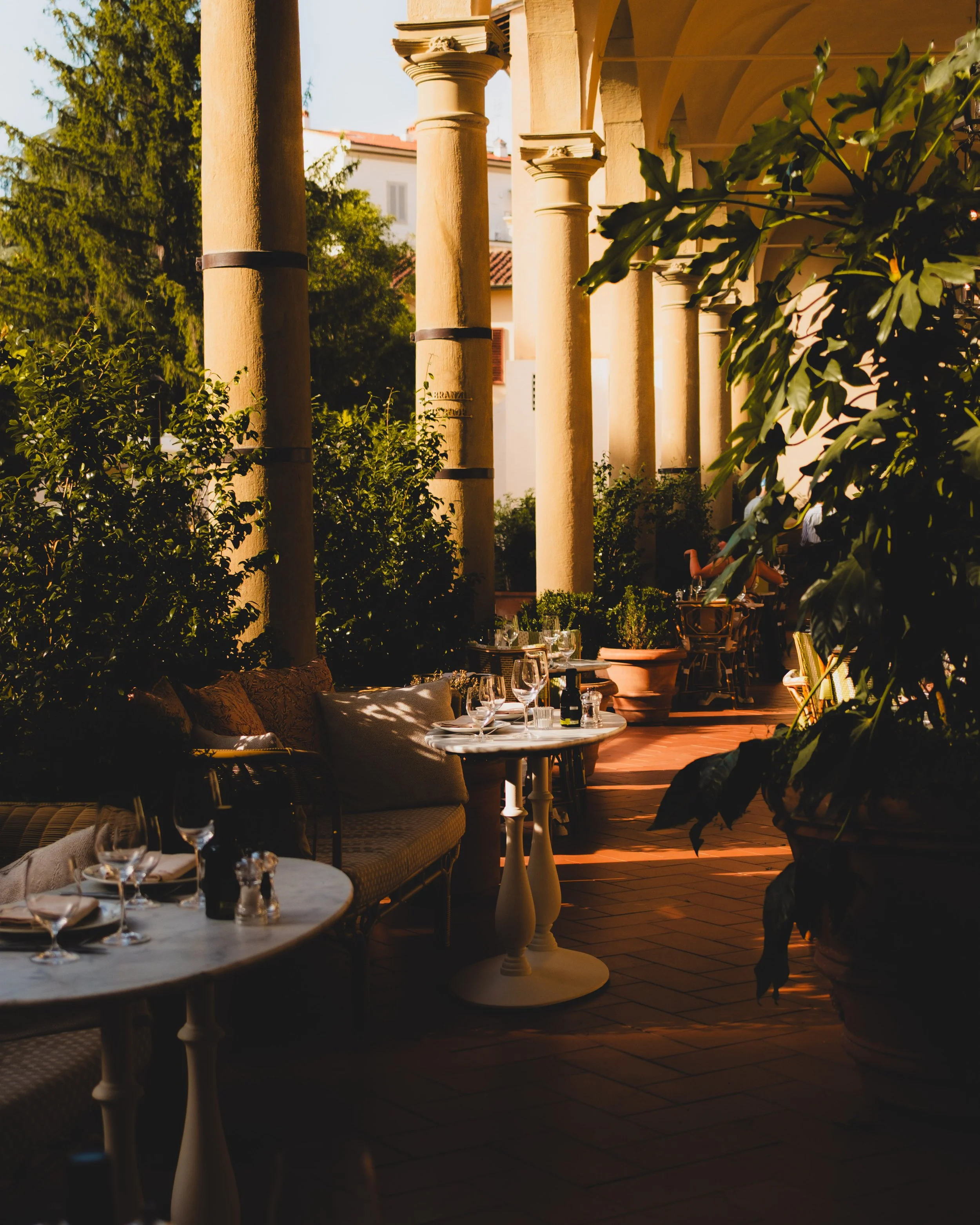 An outdoor restaurant patio with several tables set with glassware and plates, surrounded by large yellow columns and lush green plants, illuminated by warm sunlight.