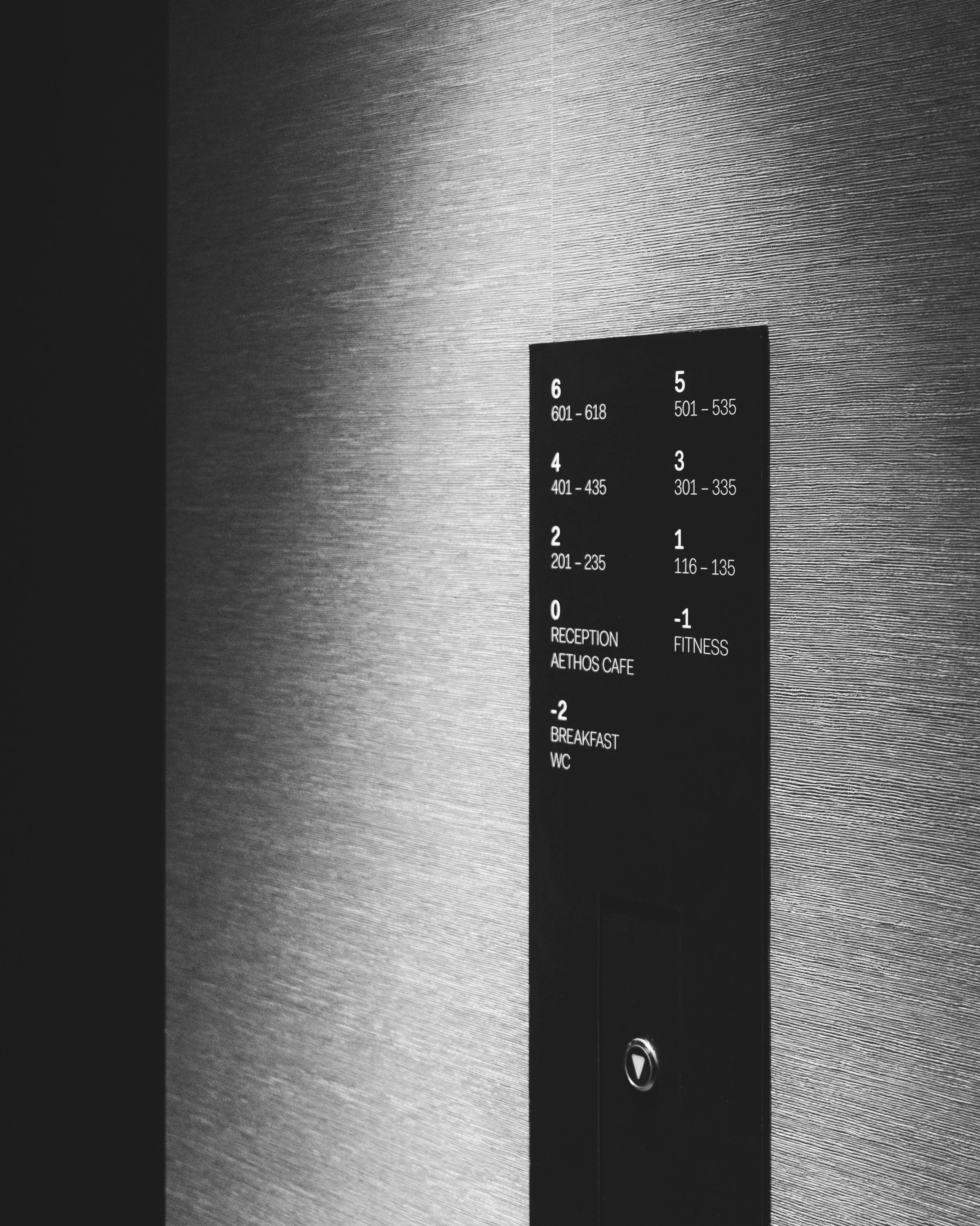 An elevator control panel on a wall with a textured surface, showing floor buttons numbered 6 through 0, with a mirror-like reflection at the bottom.