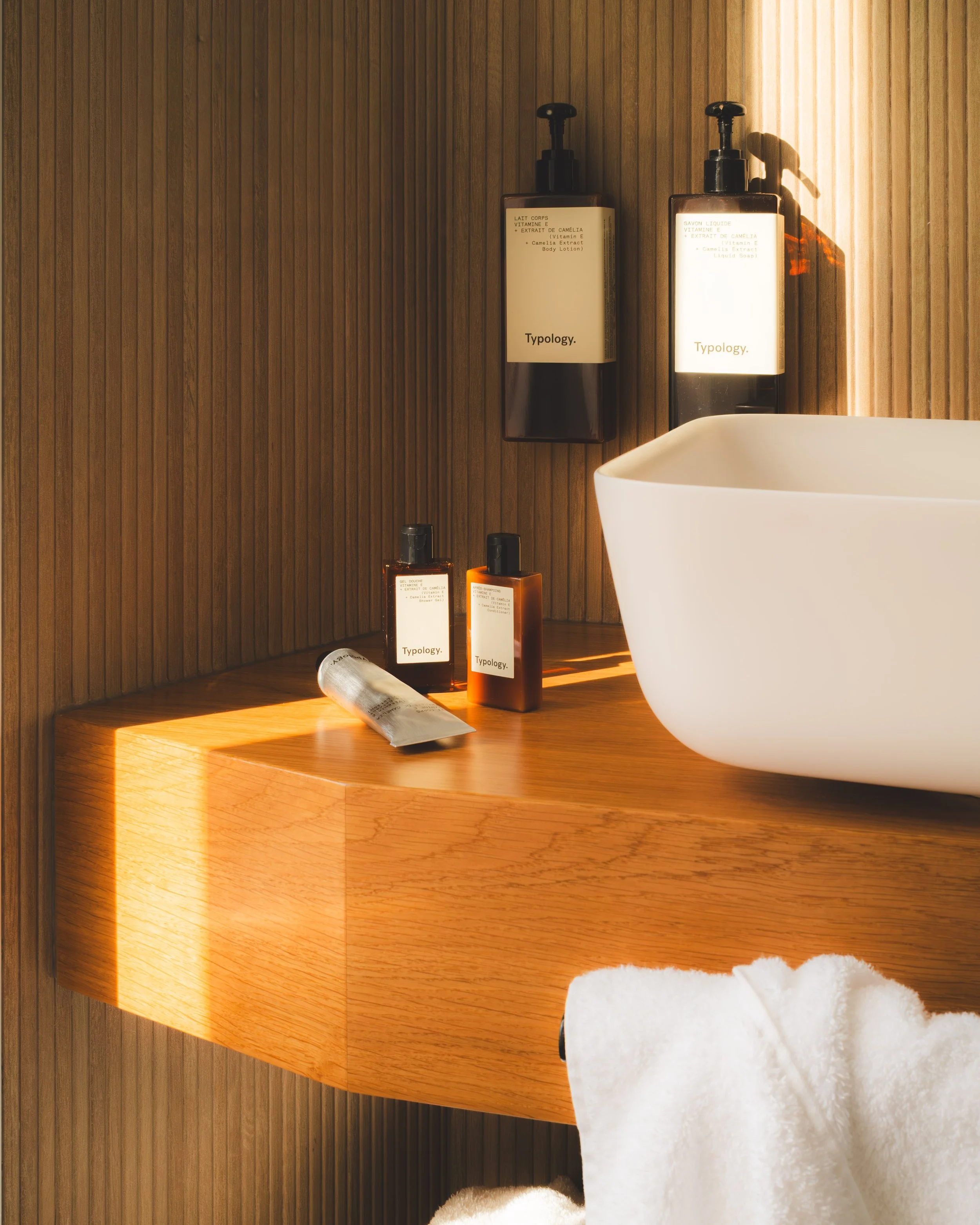 Bathroom with wooden panel walls and countertop, featuring two black pump bottles, a tube of skincare, and a white vessel sink with a fluffy towel in the foreground.