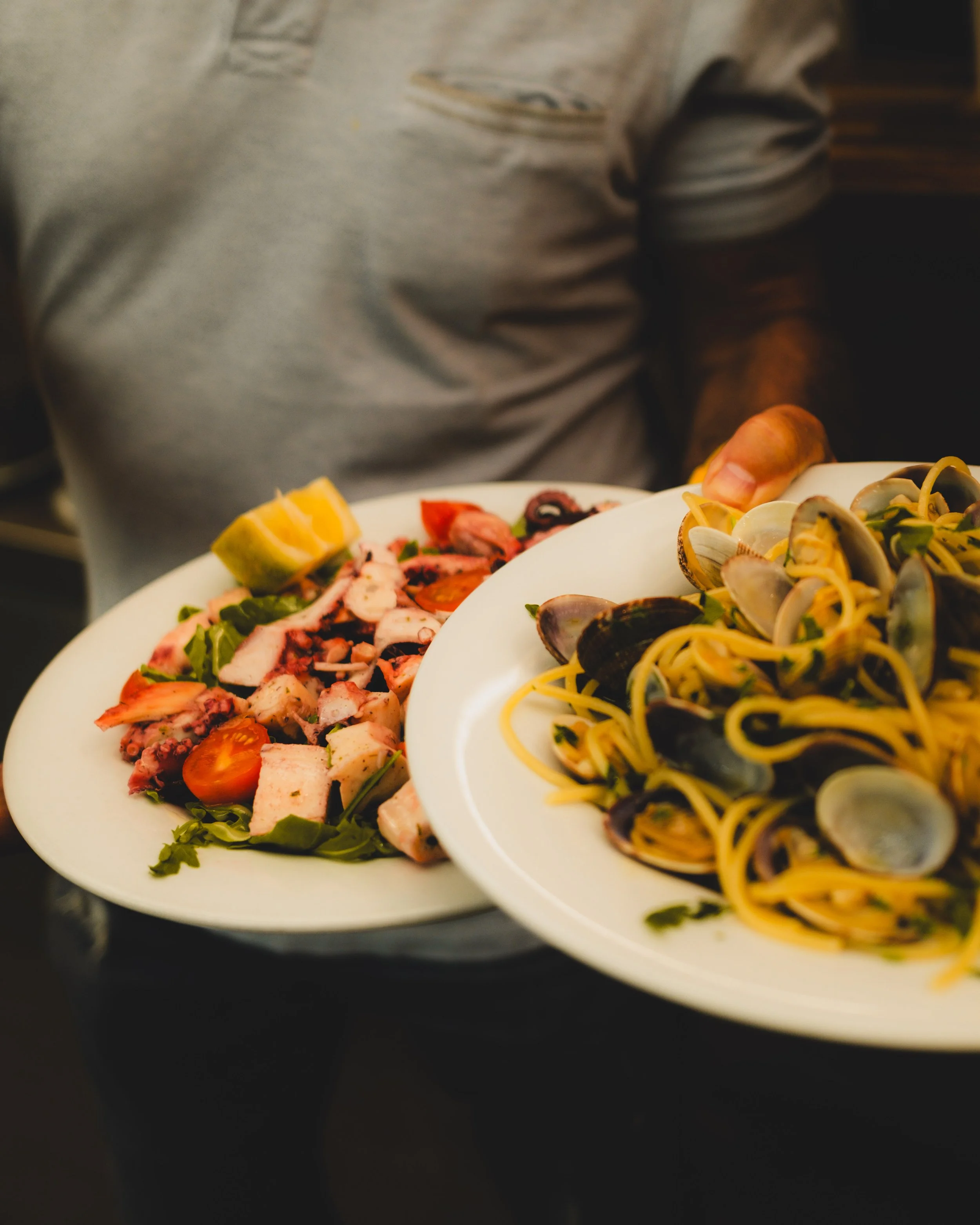 Person holding two plates of Italian food, one with seafood pasta and the other with a salad with tomatoes and grilled octopus. Lionel Chu - Hotel & Travel Photographer