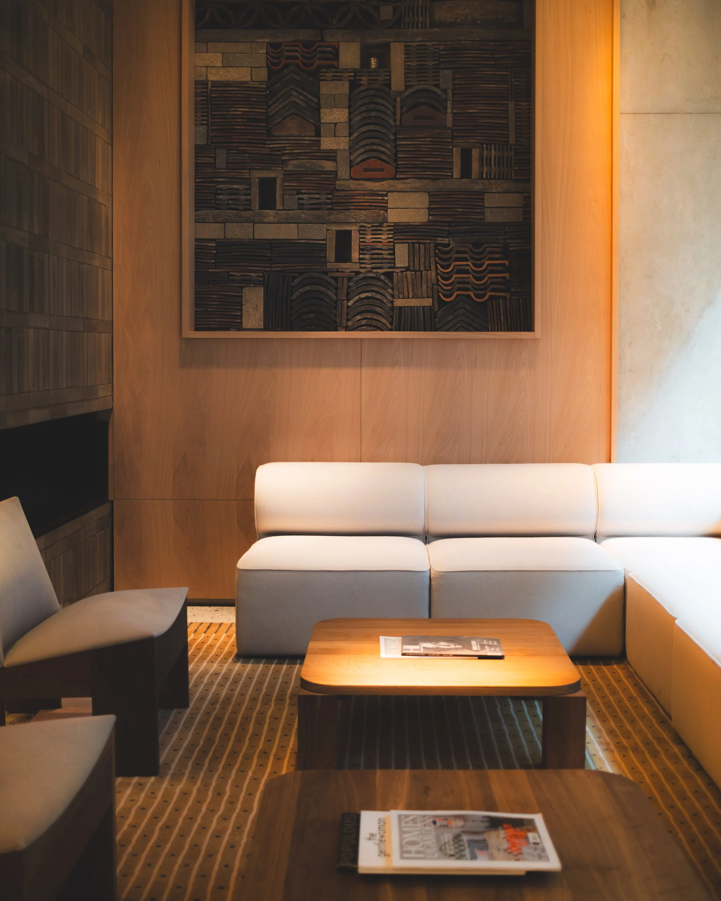 A cozy hotel lobby sitting area with a white sofa, a wooden coffee table with magazines, and wall art made of various tiles and bricks, with warm lighting and patterned carpet.