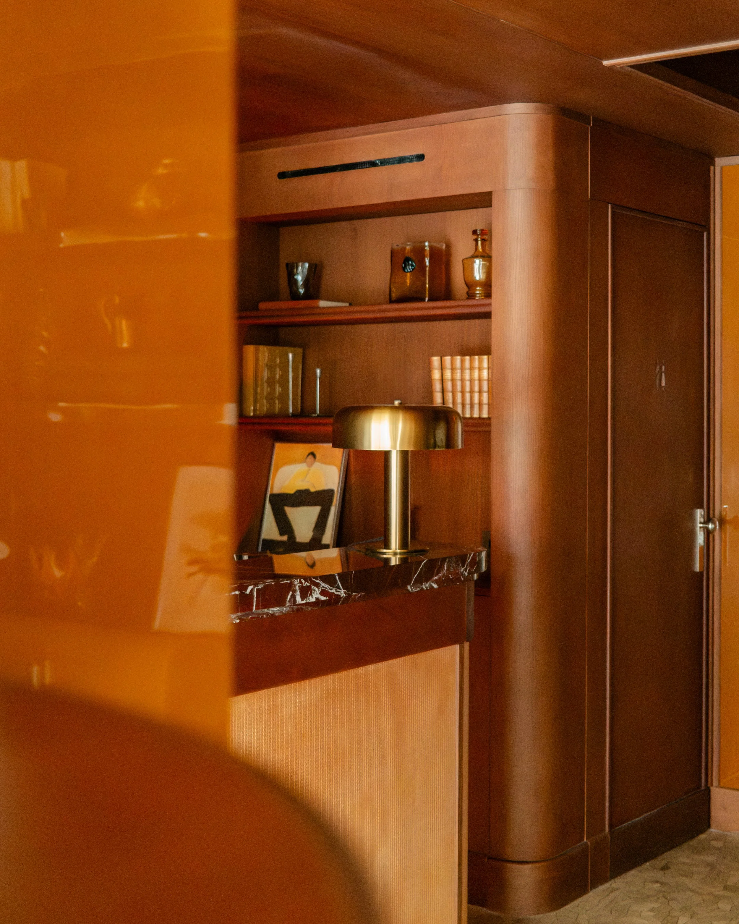 A wooden bar or reception area with a golden lamp on a dark marble top, shelves with books and decorative objects, and a framed photo leaning against the wall. Lionel Chu - Hotel & Travel Photography