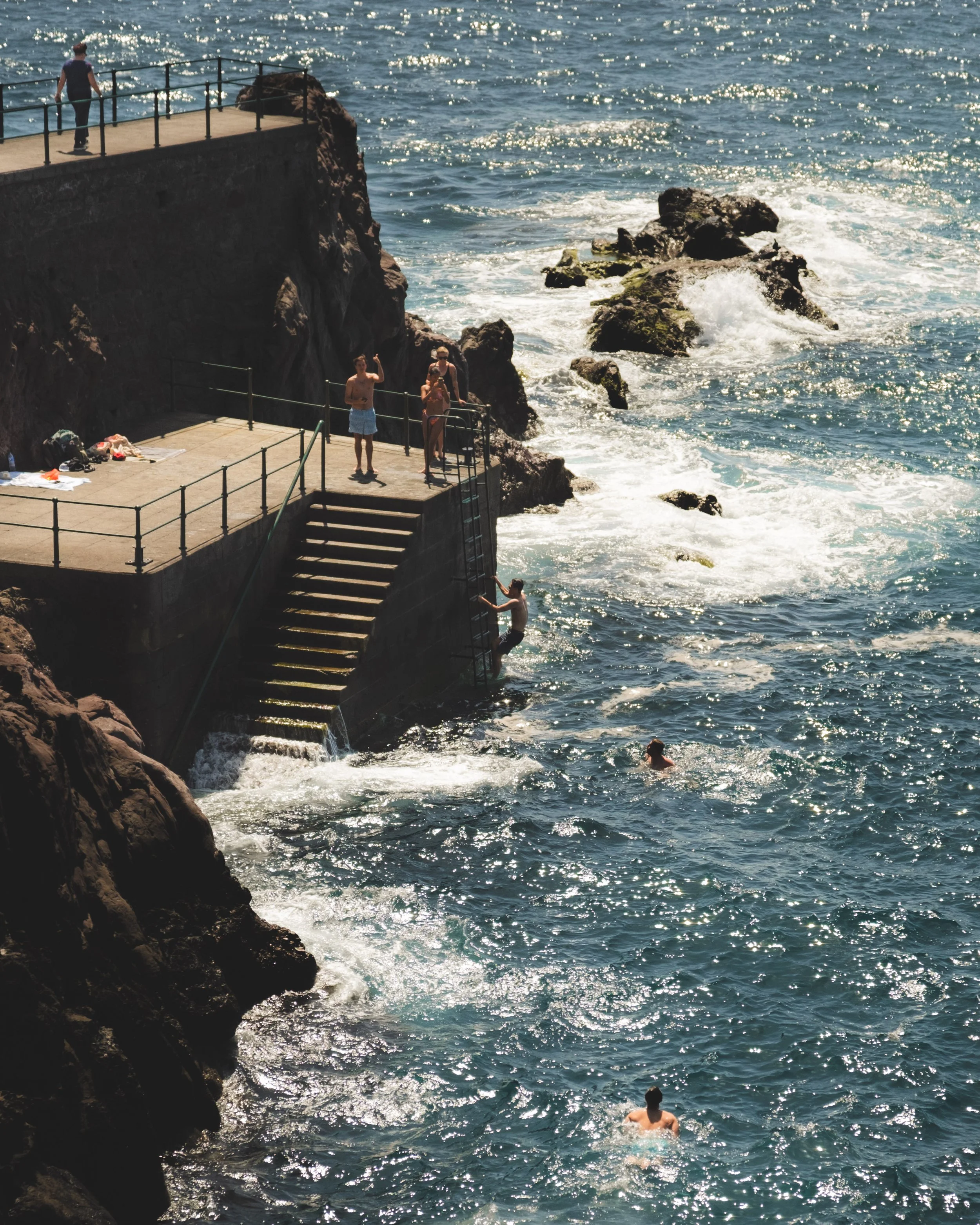 People swimming in the ocean near a rocky shoreline, with some on a concrete platform and one climbing a ladder, under bright sunlight.