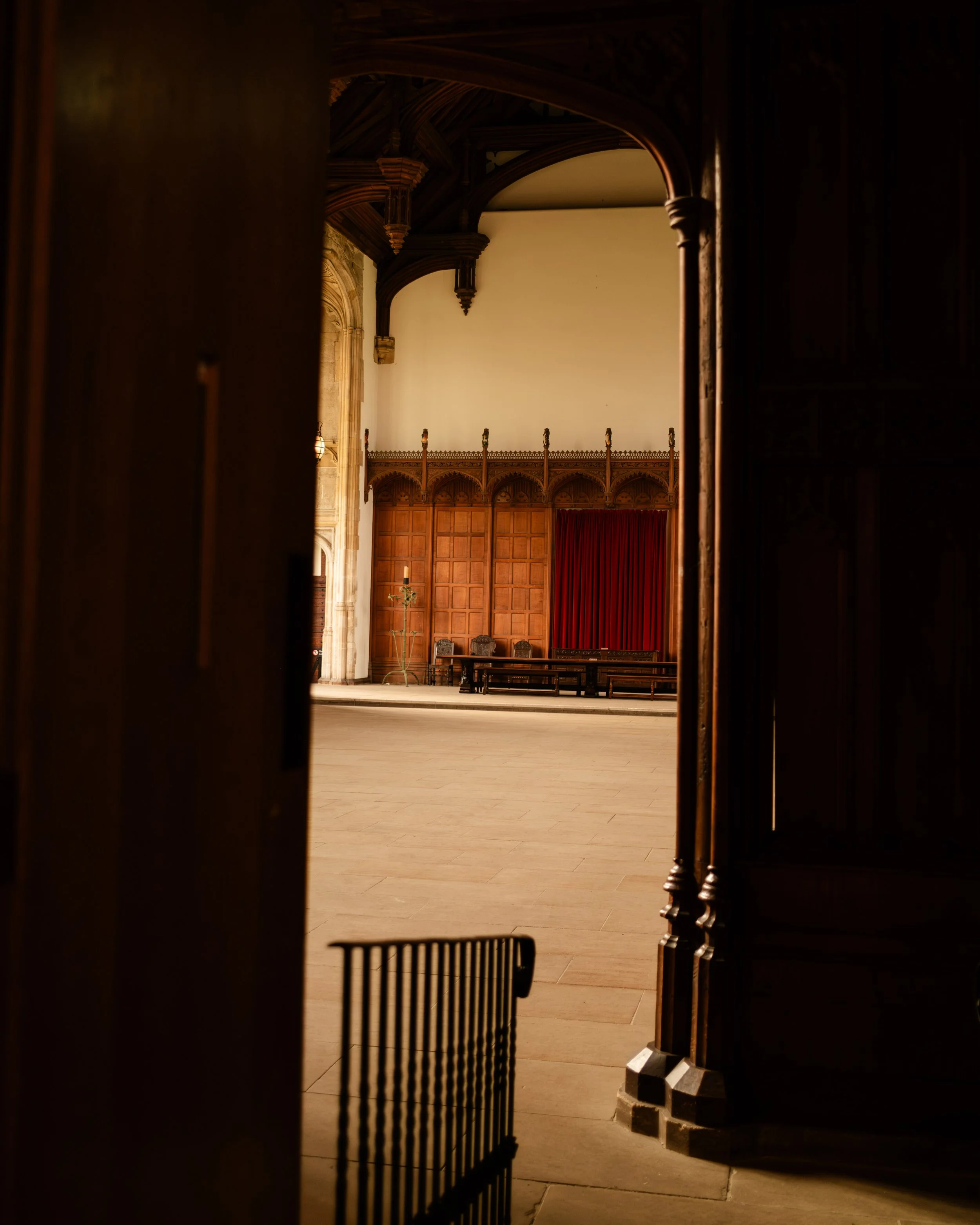 An interior view of a large room, possibly a hall or auditorium, as seen through a partially opened dark wooden door. The room features wooden paneling on the walls, a red curtain, some benches, and a decorative candelabrum, with a high ceiling suppo