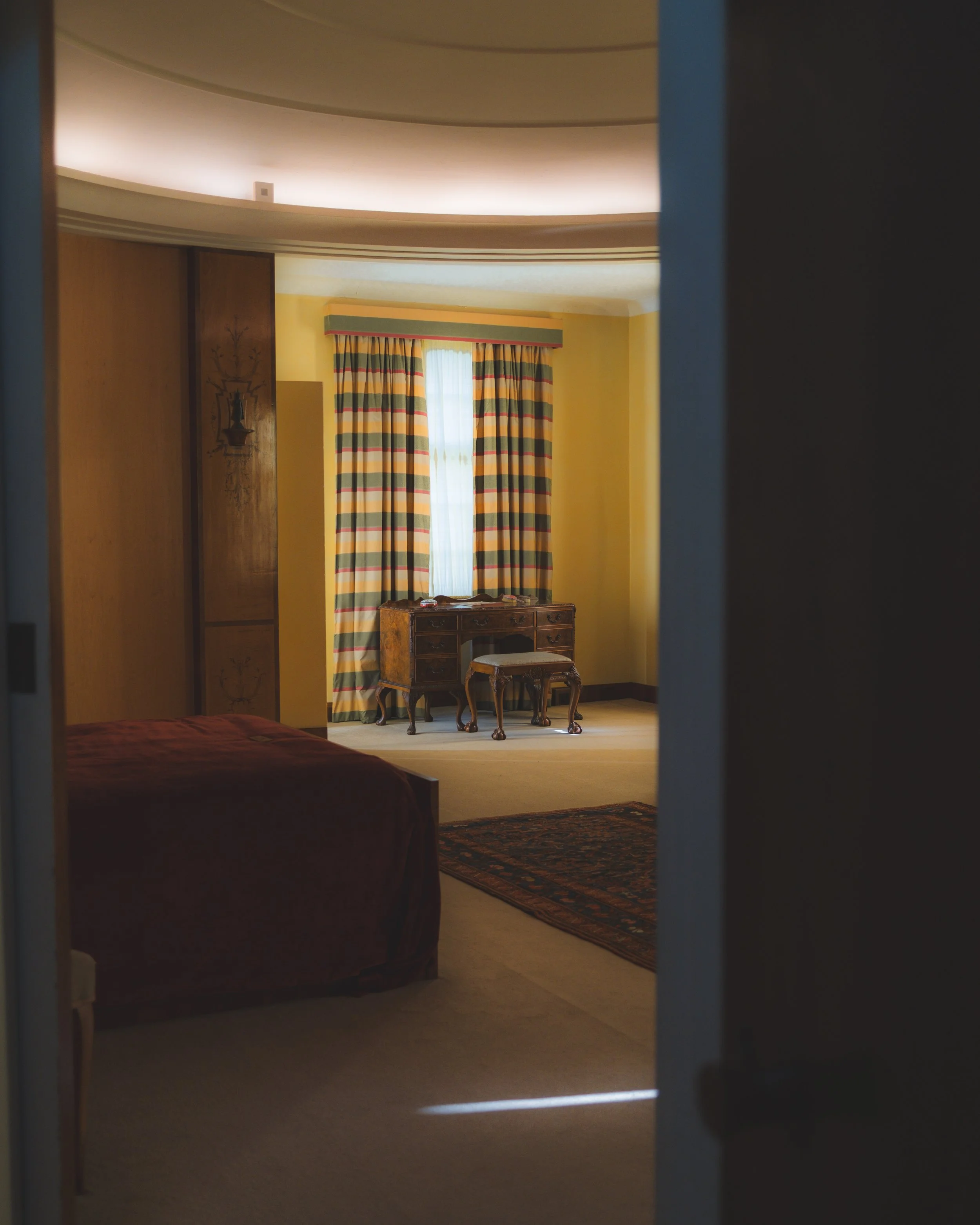 A partially visible bedroom with a yellow wall, patterned curtains, a wooden vanity table, and a chair, seen through an open doorway.