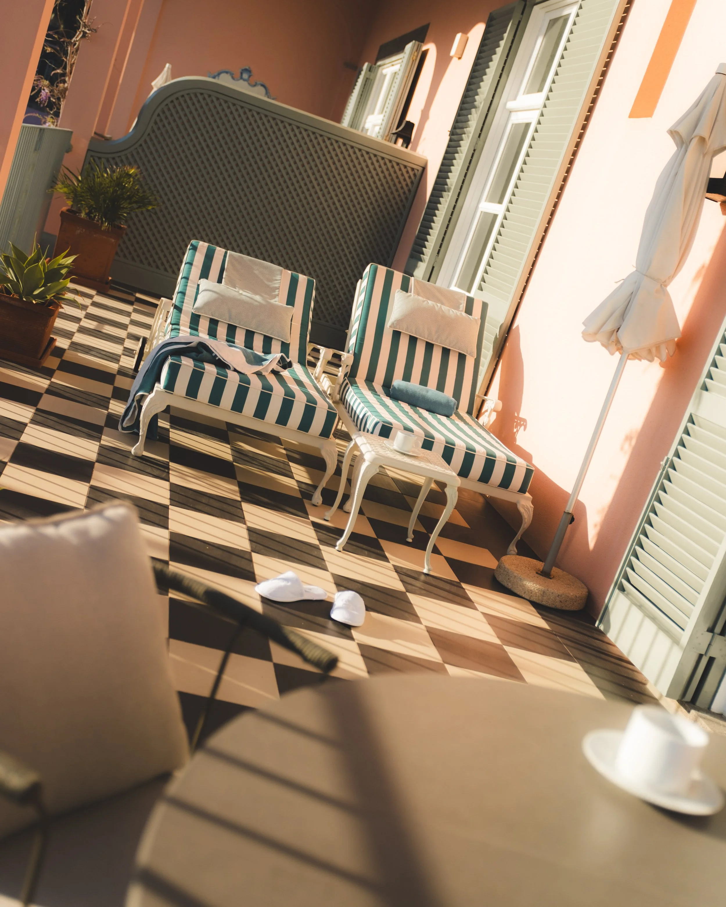Balcony with striped lounge chairs, potted plants, and a white umbrella next to a wall with windows and shutters, sunlight casting shadows.