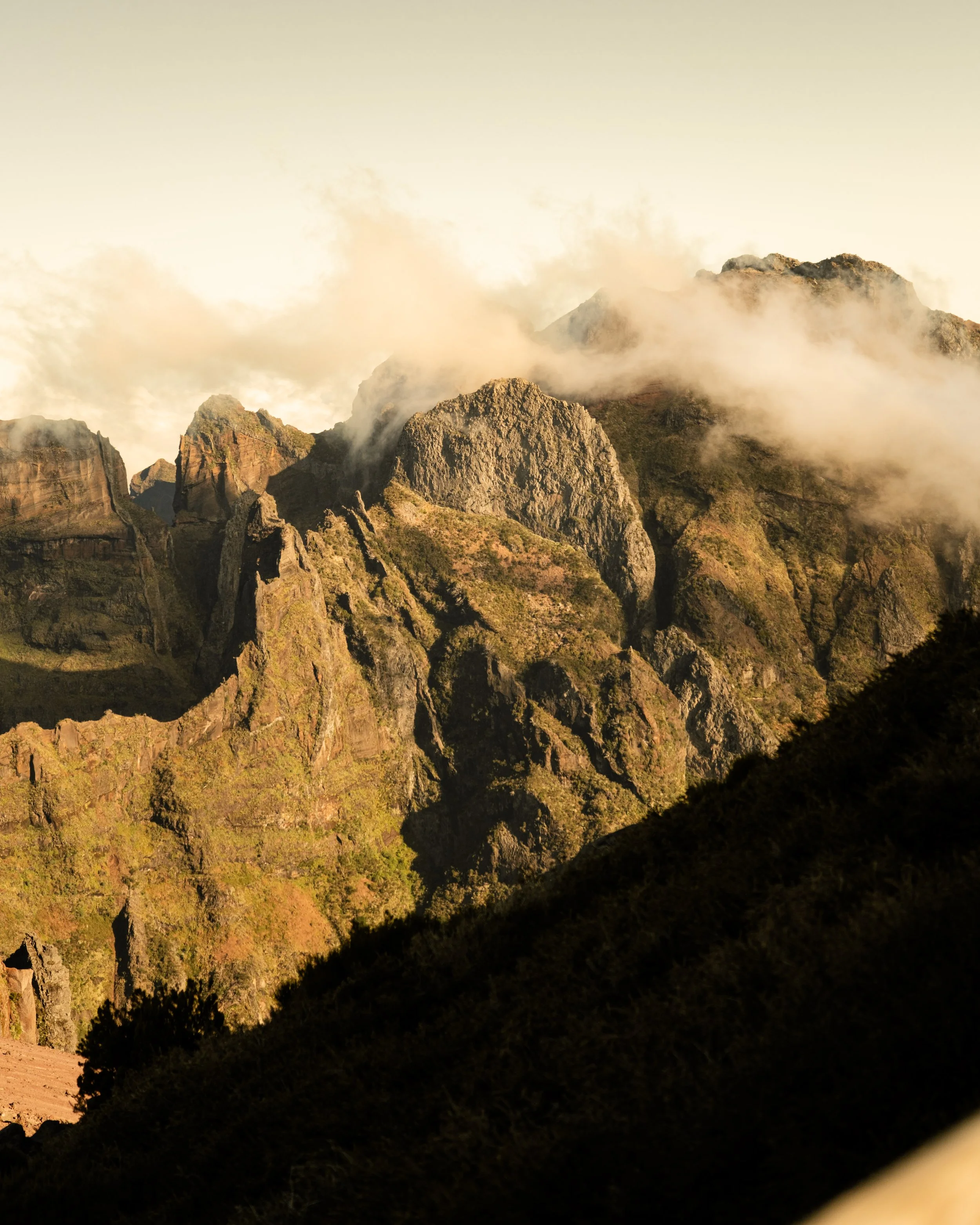 Sunlit rugged mountain range with mist and clouds around the peaks, viewed during daylight hours.